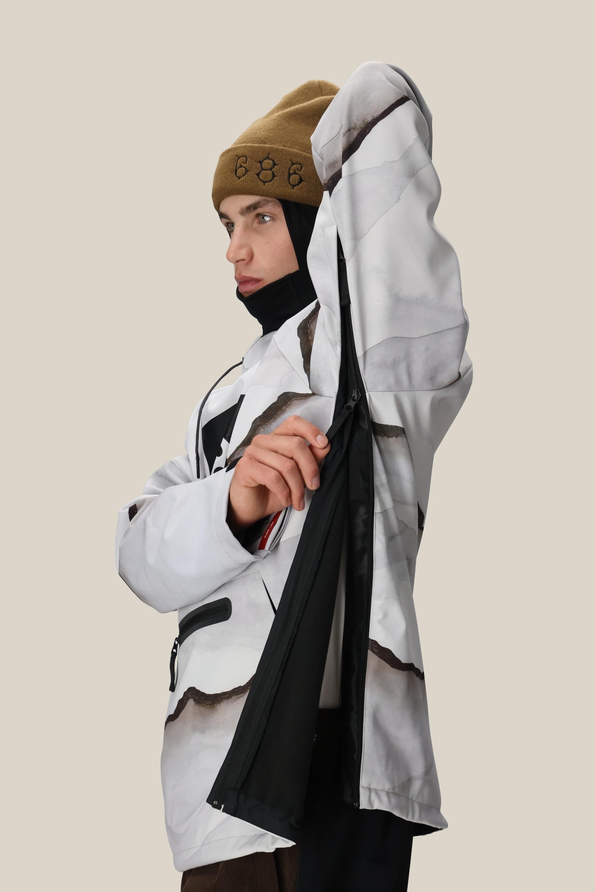 A person wearing the 686 Mens Waterproof Hoody by 686 opens a large side zipper to reveal its fleece-lined inner mesh, while looking to the side against a plain beige background.