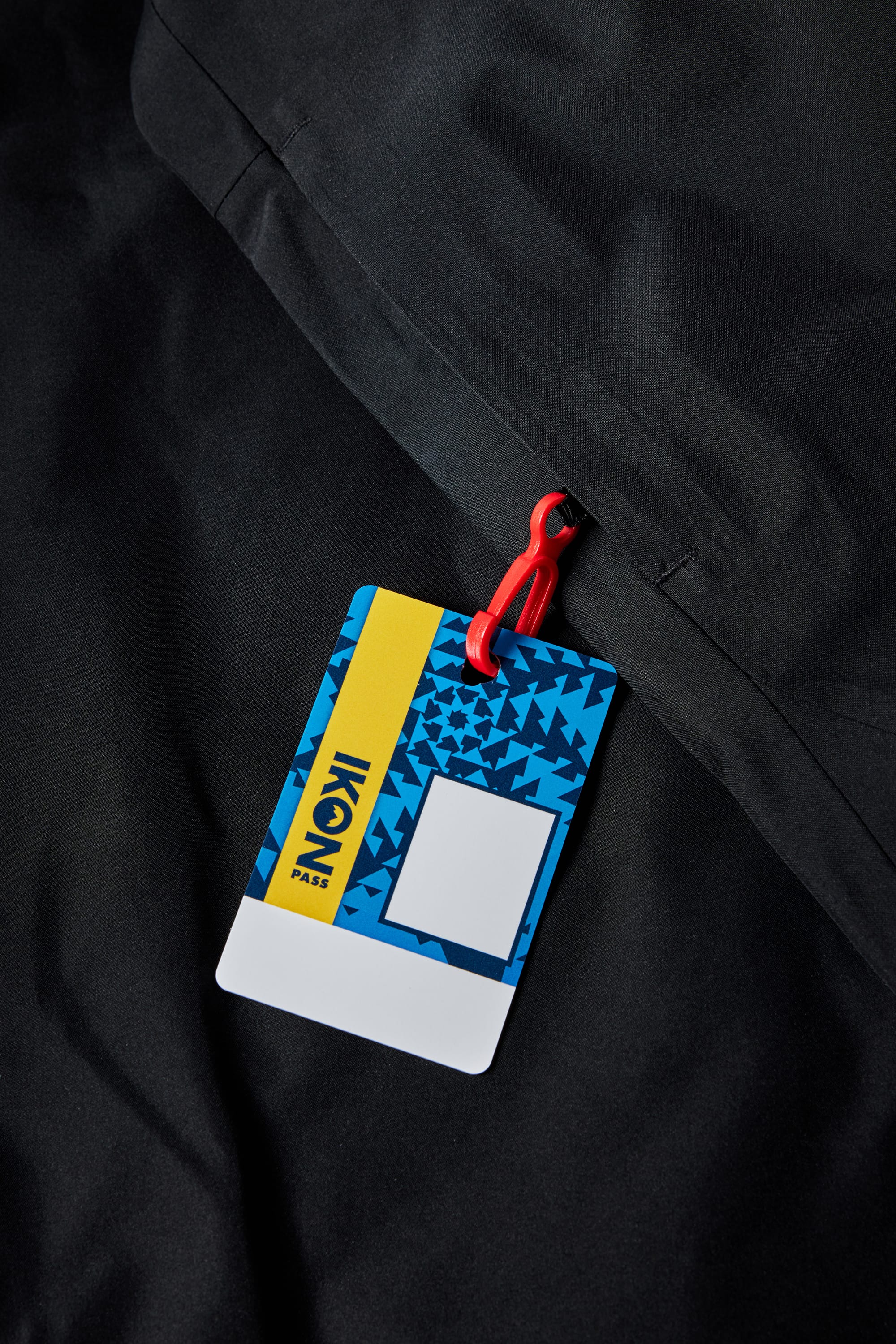 A colorful Ikon Pass ski lift ticket with a red clip is attached to the zipper of the black 686 Mens Waterproof Hoody by 686, featuring a fleece lining. The pass displays a blue geometric design, yellow stripe, and space for labeling.