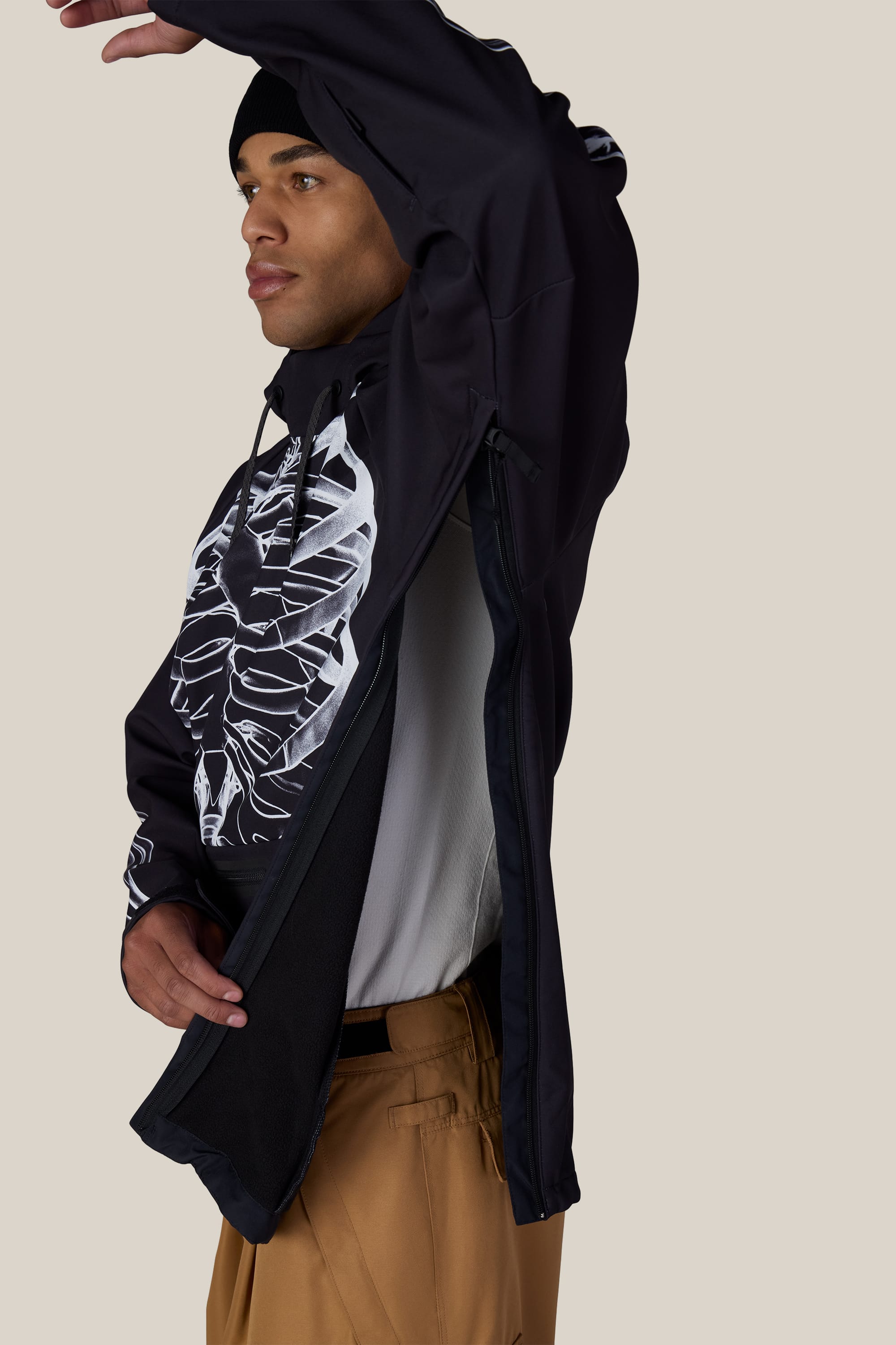 A person wearing the 686 Men’s Waterproof Hoody in tan pants lifts their arm, revealing the side zipper and mesh lining of the black jacket with a white abstract design by 686. The background is plain beige.