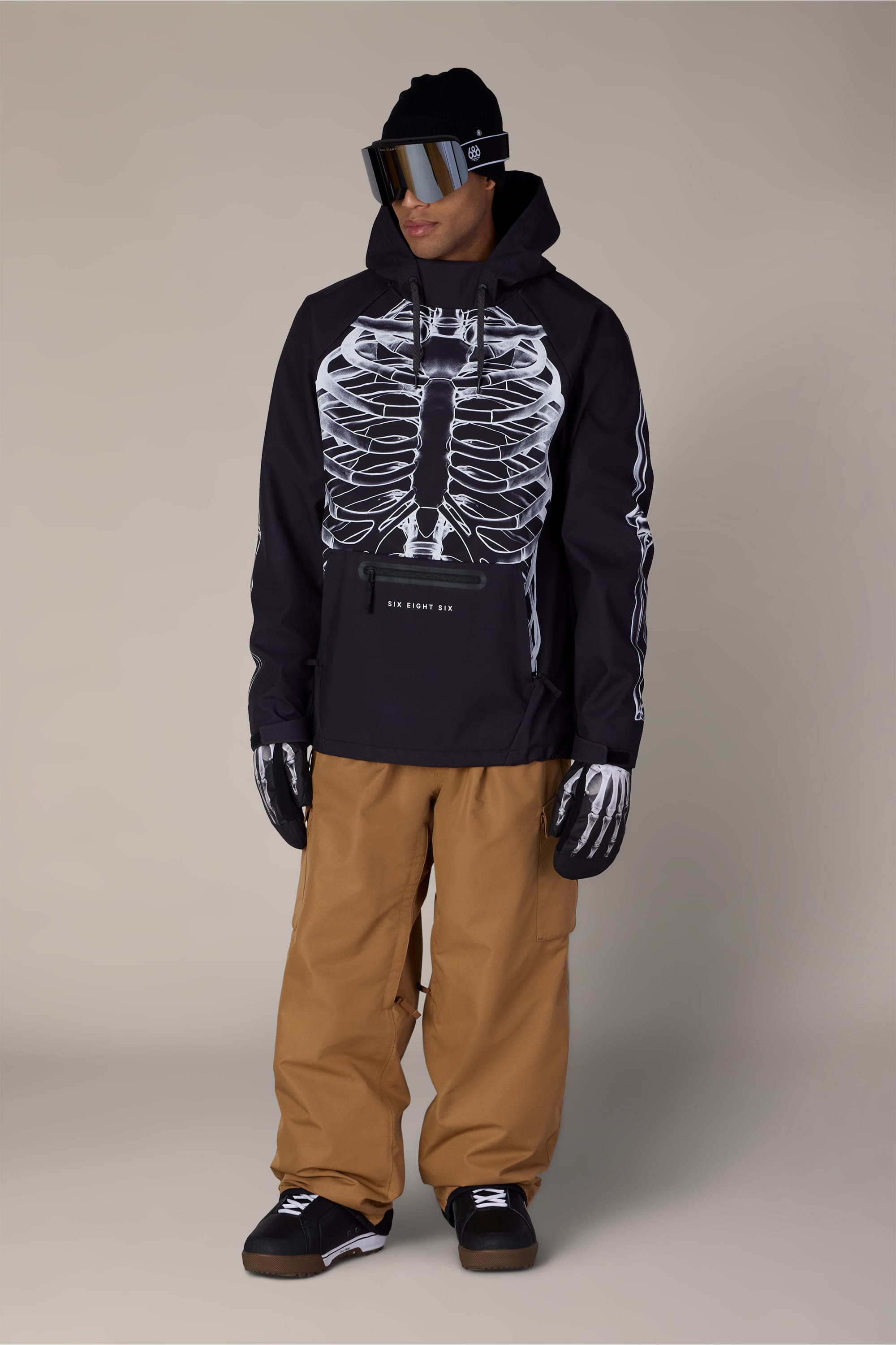 A person wearing the 686 Men’s Waterproof Hoody in black, featuring a white skeleton print, paired with tan snow pants, black gloves with skeleton prints, black winter boots, ski goggles, and a black beanie stands against a plain background.