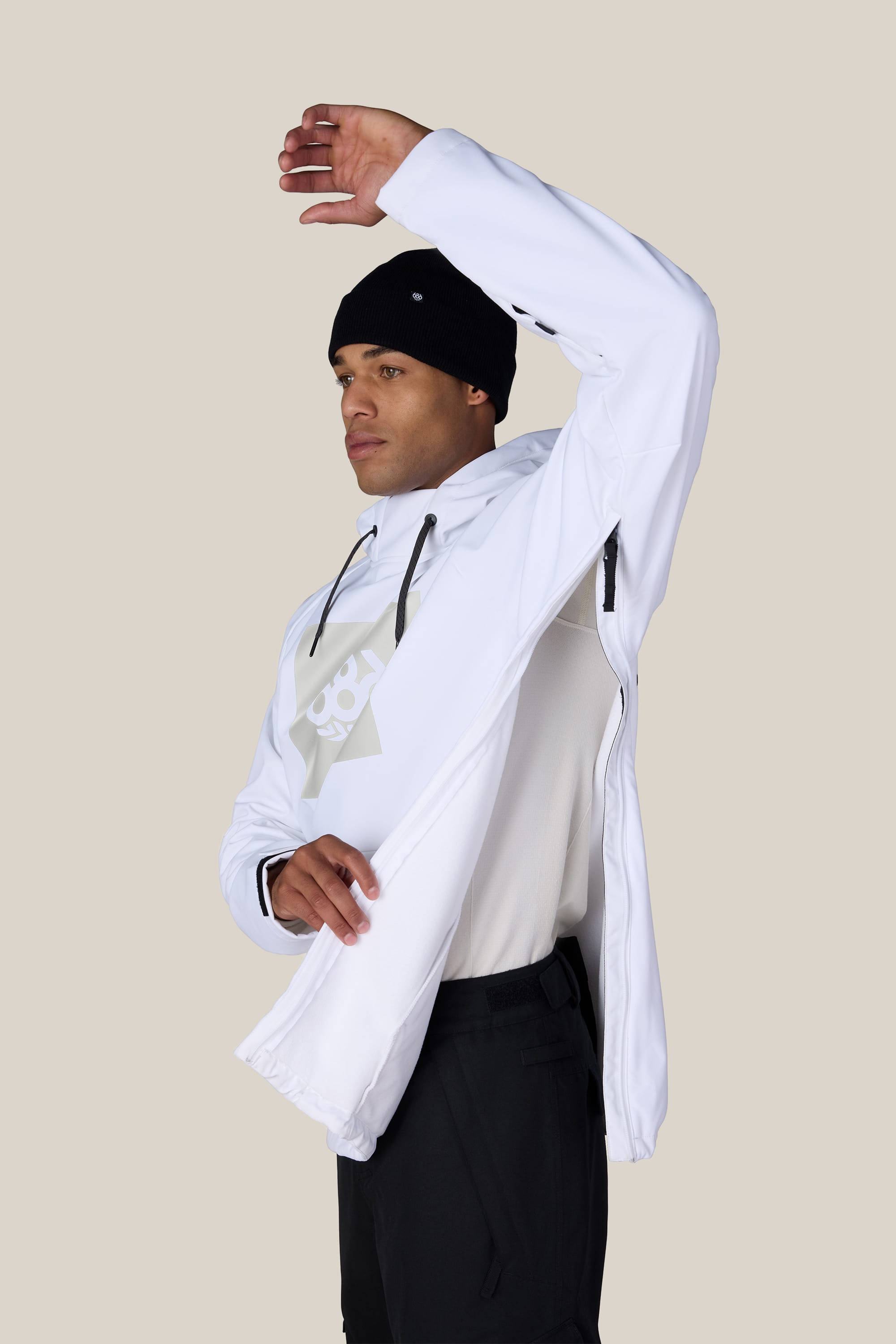 A person wearing the 686 Men’s Waterproof Hoody by 686, along with a white shirt, black pants, and a black beanie, raises one arm to zip up the side of their hoody against a neutral background.