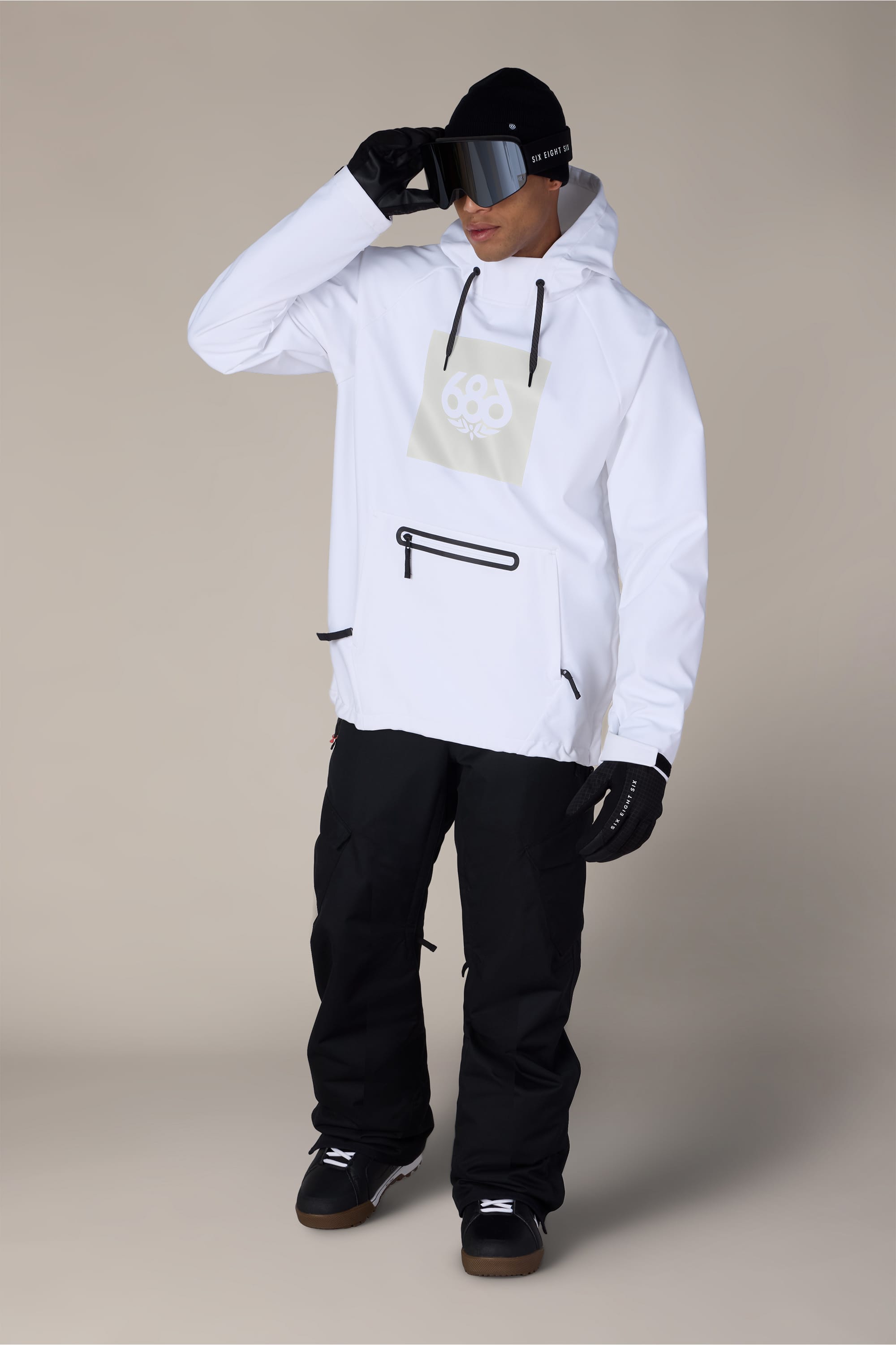 A person wearing the 686 Men’s Waterproof Hoody, black snow pants, gloves, and ski goggles stands against a plain background, holding the goggles with one hand.