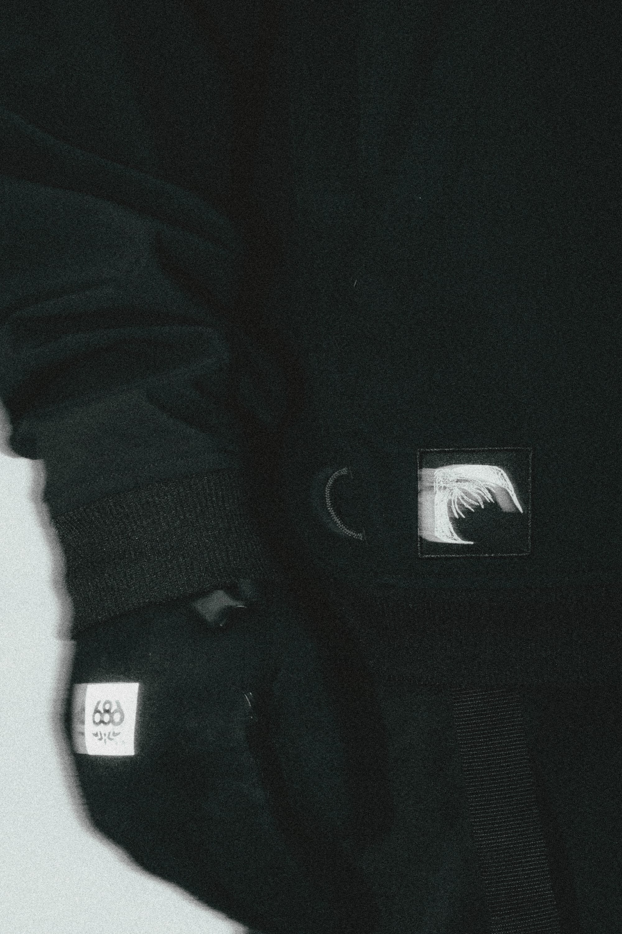 A person wears the 686 Mens Waterproof Team Hoody—dark, long-sleeved, with a hand in the secure pocket. Visible: a circular 686 logo patch on the sleeve and a rectangular patch with a stylized wave or feather near the waist.