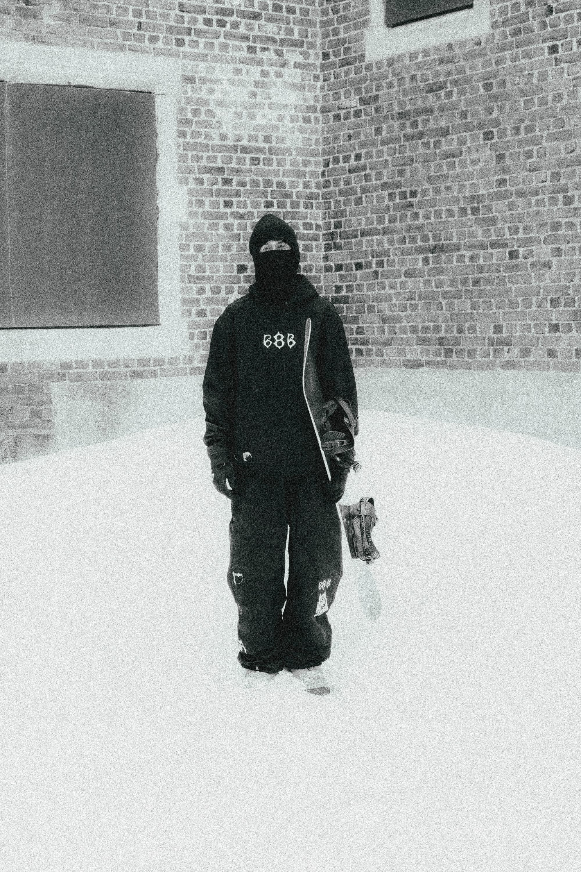 Wearing the 686 Mens Waterproof Team Hoody and a balaclava, a person stands on snow with a snowboard before a brick building. The high-contrast, grainy, black-and-white photo highlights the 686 brand style.