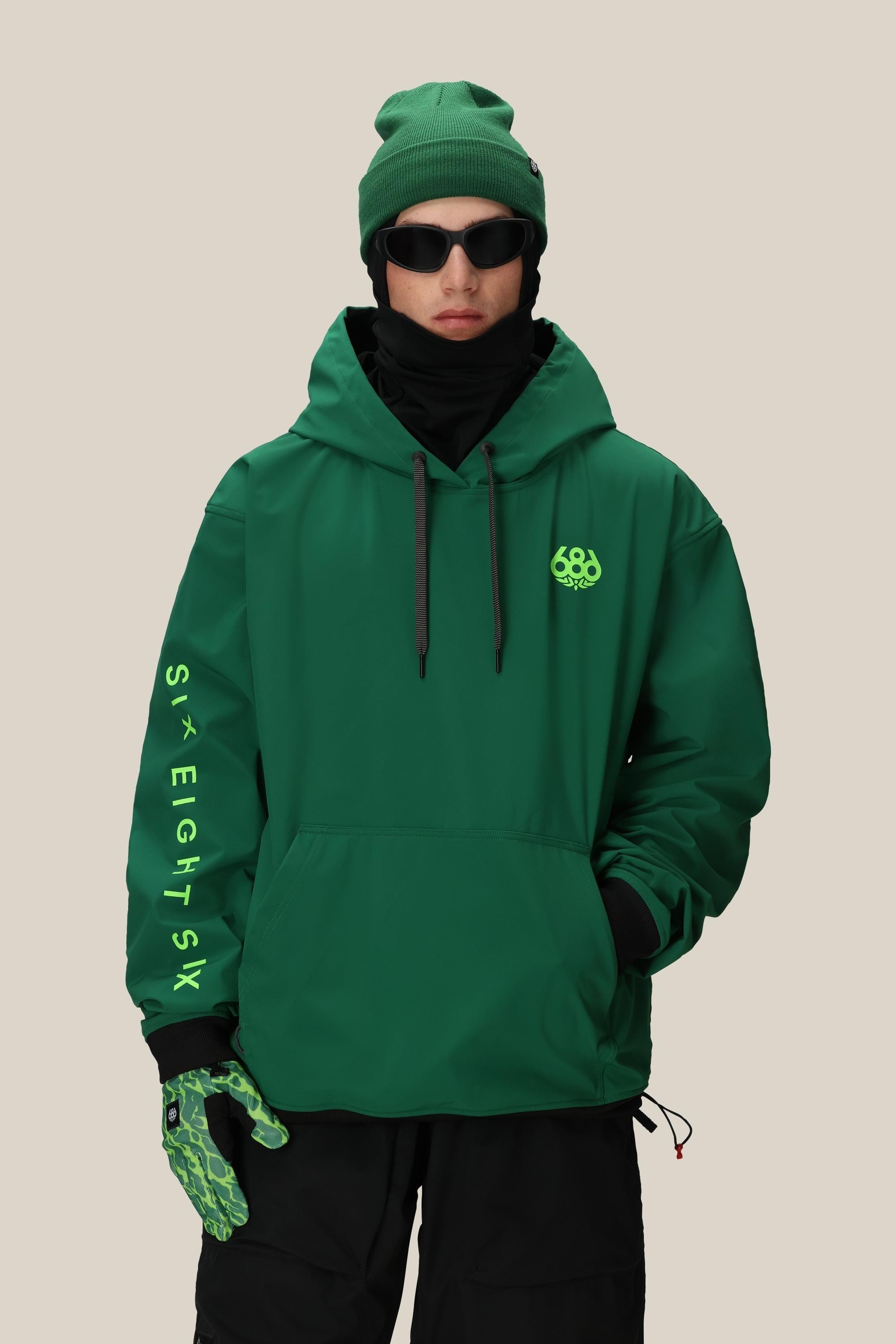 Wearing the 686 Mens Waterproof Team Hoody in green with neon green 686 and SIX EIGHT SIX prints, plus a matching green beanie, black gloves with green accents, black sunglasses, and black pants, a person poses against a beige background.
