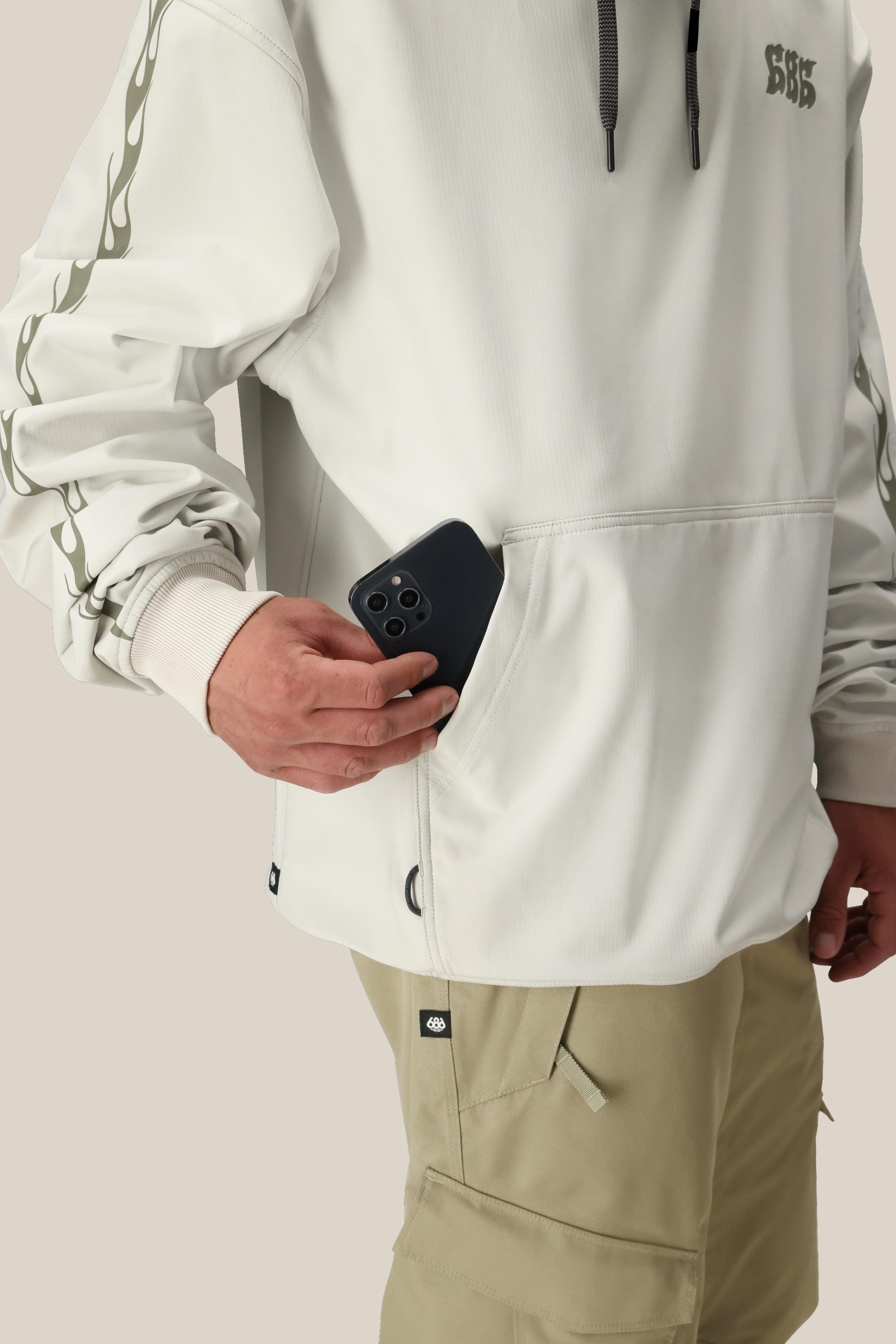 A person wearing the 686 Mens Waterproof Team Hoody with flame sleeve designs and beige cargo shorts is placing a black smartphone into the front pocket. The plain, neutral background highlights the stylish 686 hoody.