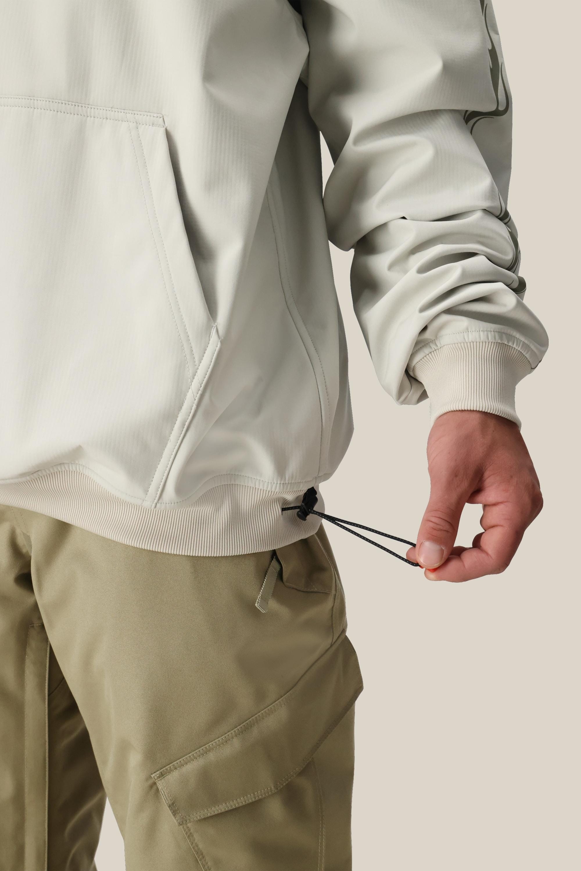 A person wearing the 686 Mens Waterproof Team Hoody in a light color with khaki cargo pants adjusts the hoodys hem drawstring. The background is plain light beige.
