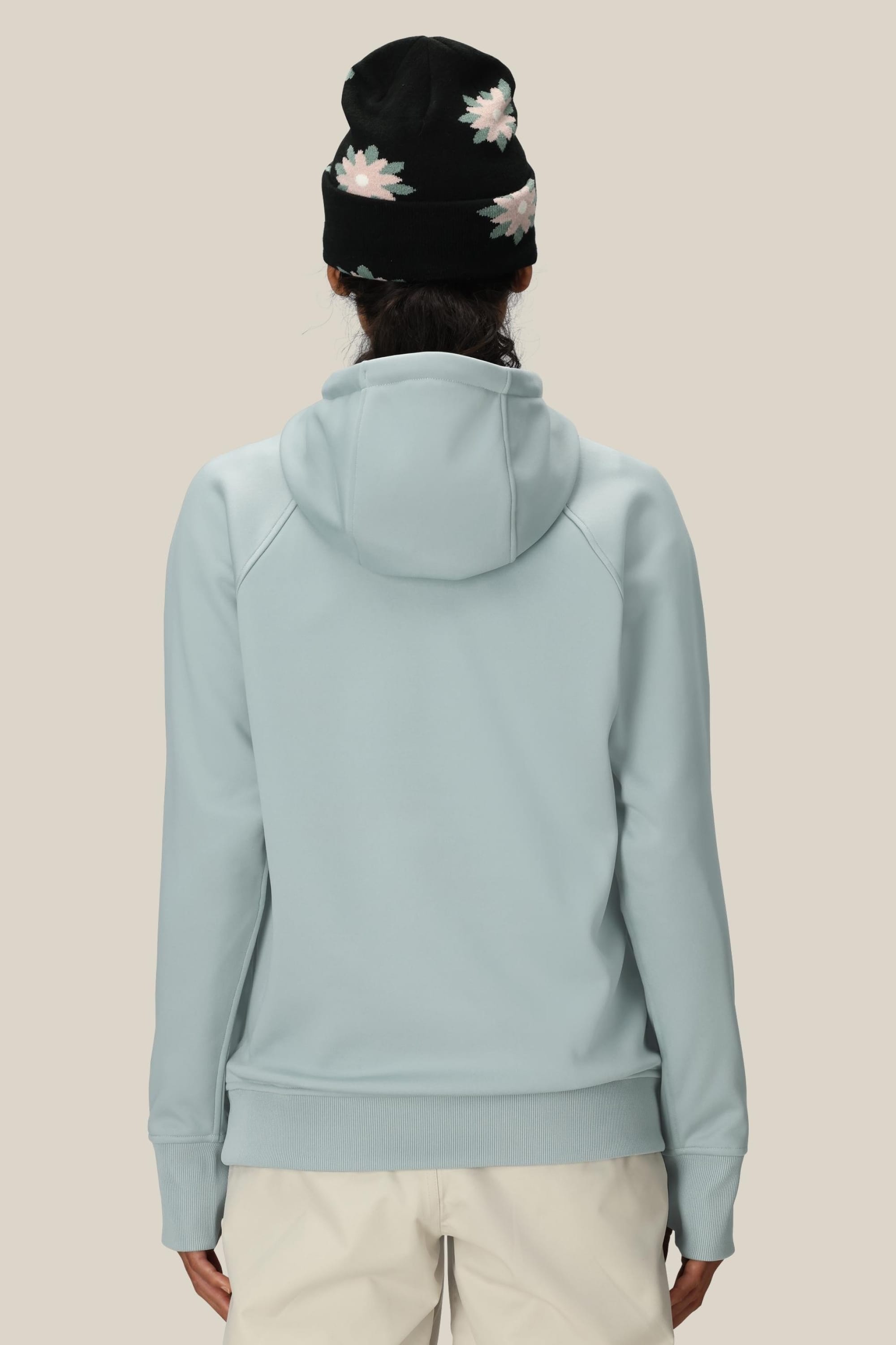 A person stands with their back to the camera, wearing the 686 Womens Bonded Fleece Pullover Hoody in light blue and a black beanie with white floral patterns, set against a plain beige background.