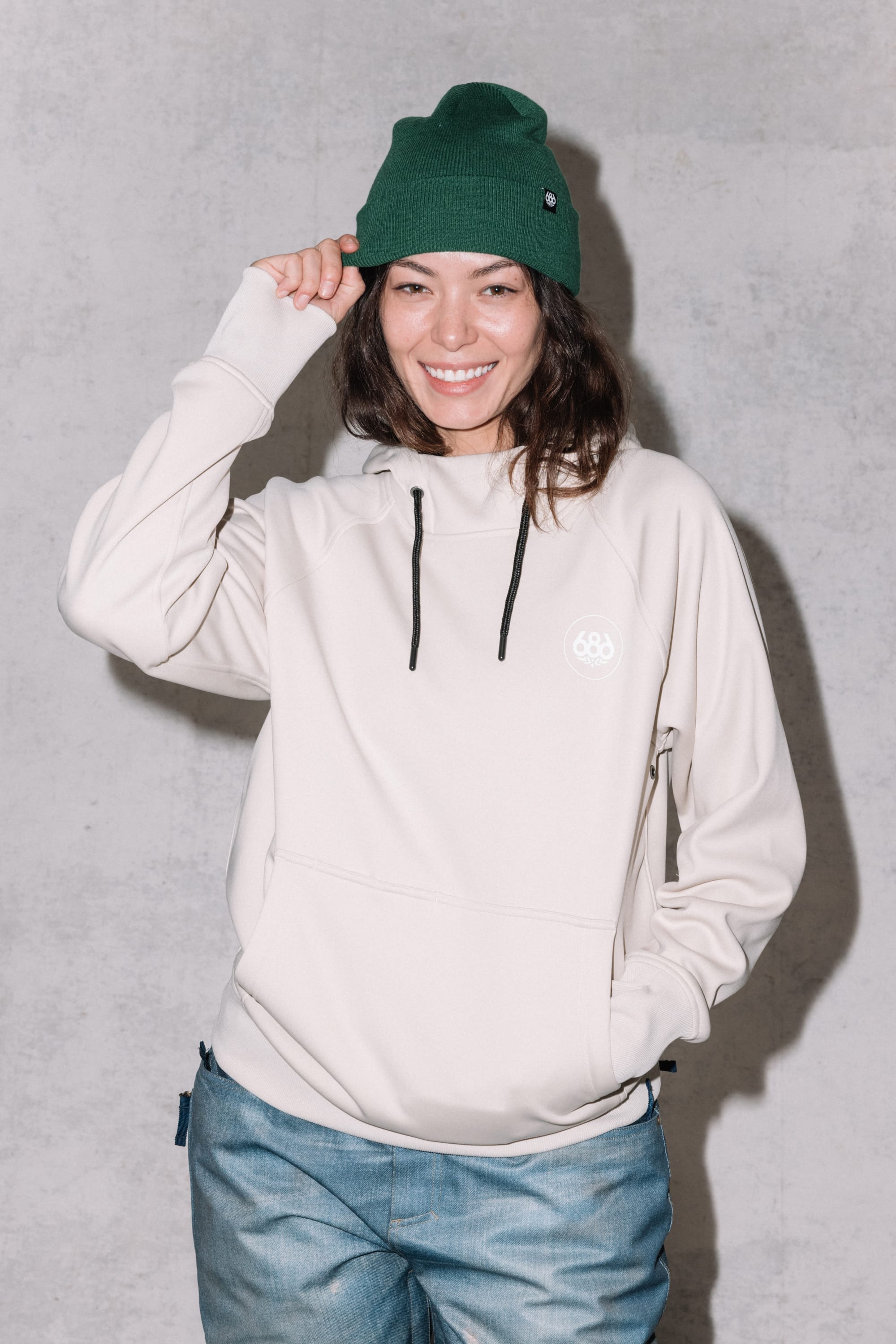 A smiling woman wears the 686 Womens Bonded Fleece Pullover Hoody, blue jeans, and a green knit beanie while standing against a light gray background, touching the brim of her beanie with one hand.