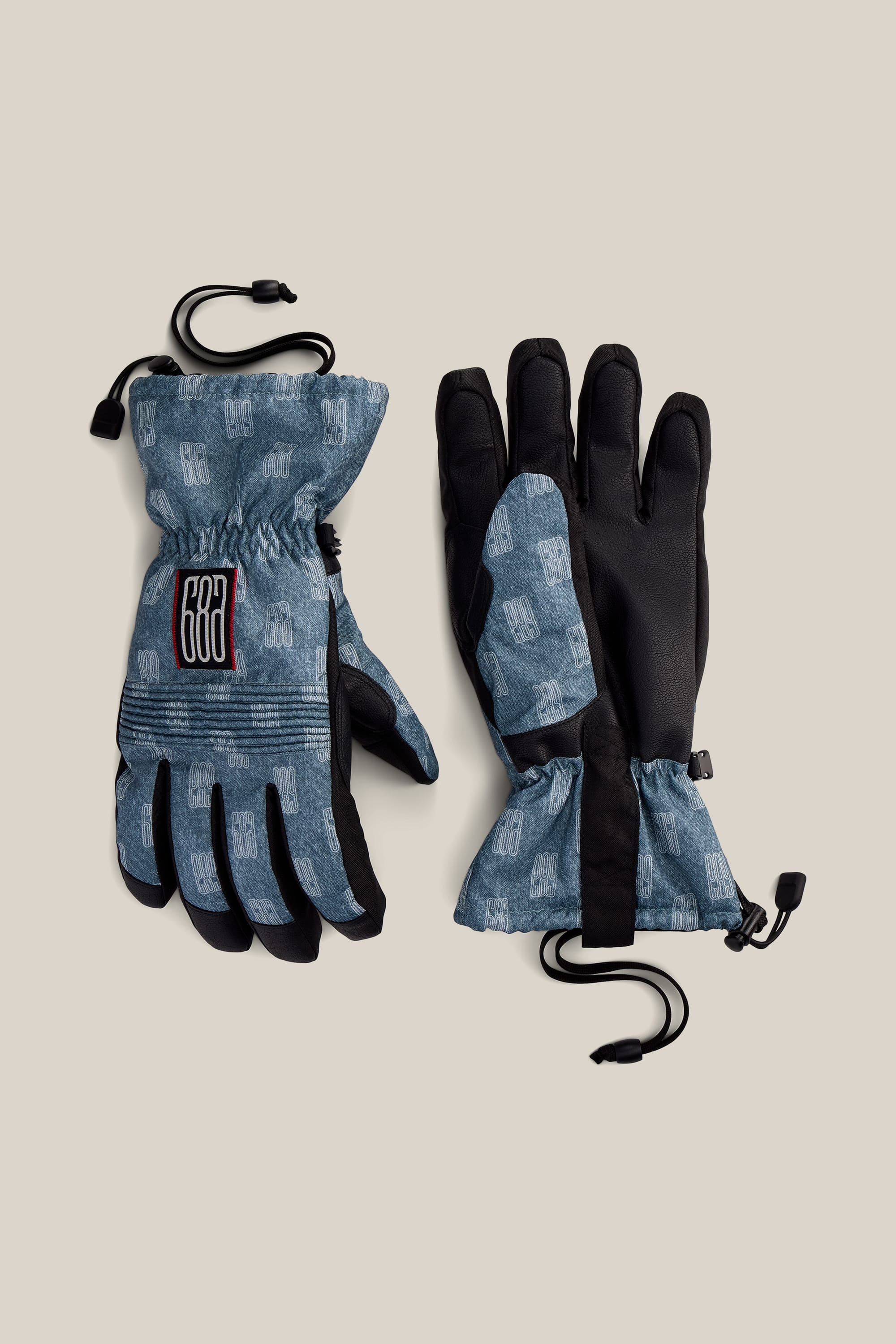 A pair of 686 Men’s Lander™ Gloves in blue and black, featuring adjustable wrist straps and a geometric pattern, displayed on a beige background.