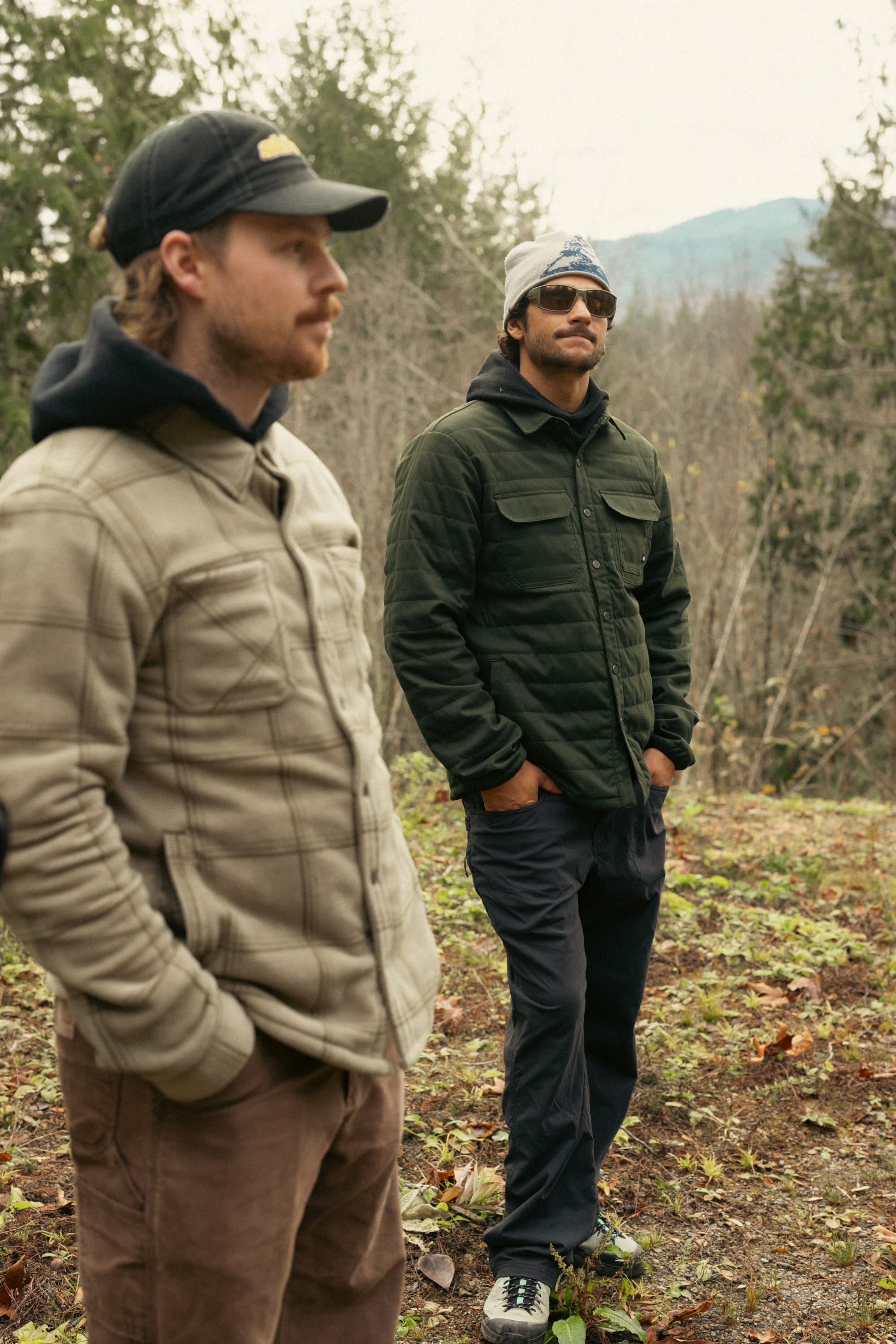 Two men stand outdoors amid trees and mountains. One wears the beige 686 Men's Thermaldry™ Snap-Up Shirt with a cap and beard; the other has dark green citywear, sunglasses, and a beanie.