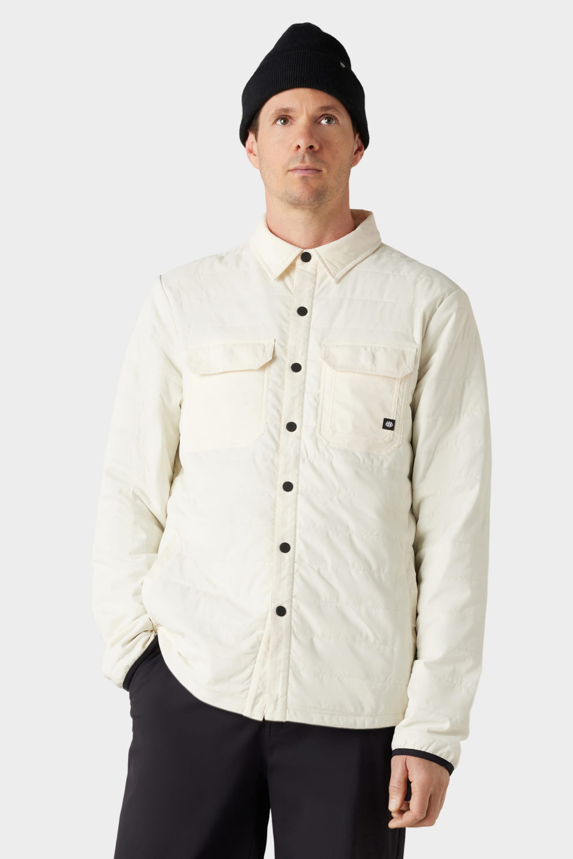 A man wears the 686 Men's Thermaldry™ Snap-Up Shirt in off-white, featuring chest pockets, paired with a black beanie and black pants, standing against a plain light gray background.