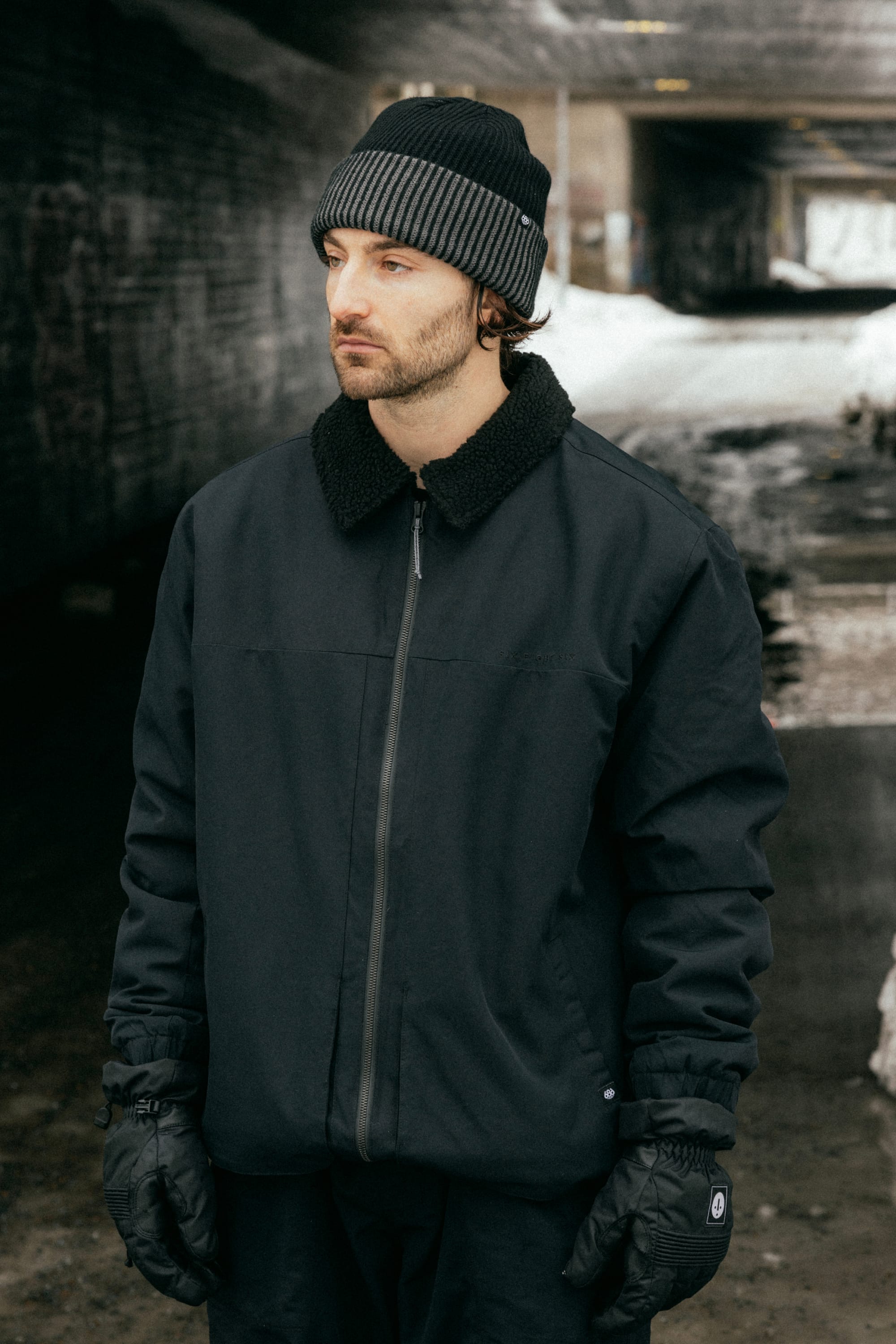 A man wearing the 686 Mens Caim Insulated Jacket stands outdoors in a snowy urban setting, paired with black gloves and a gray-striped black knit hat, looking to the side with a neutral expression.