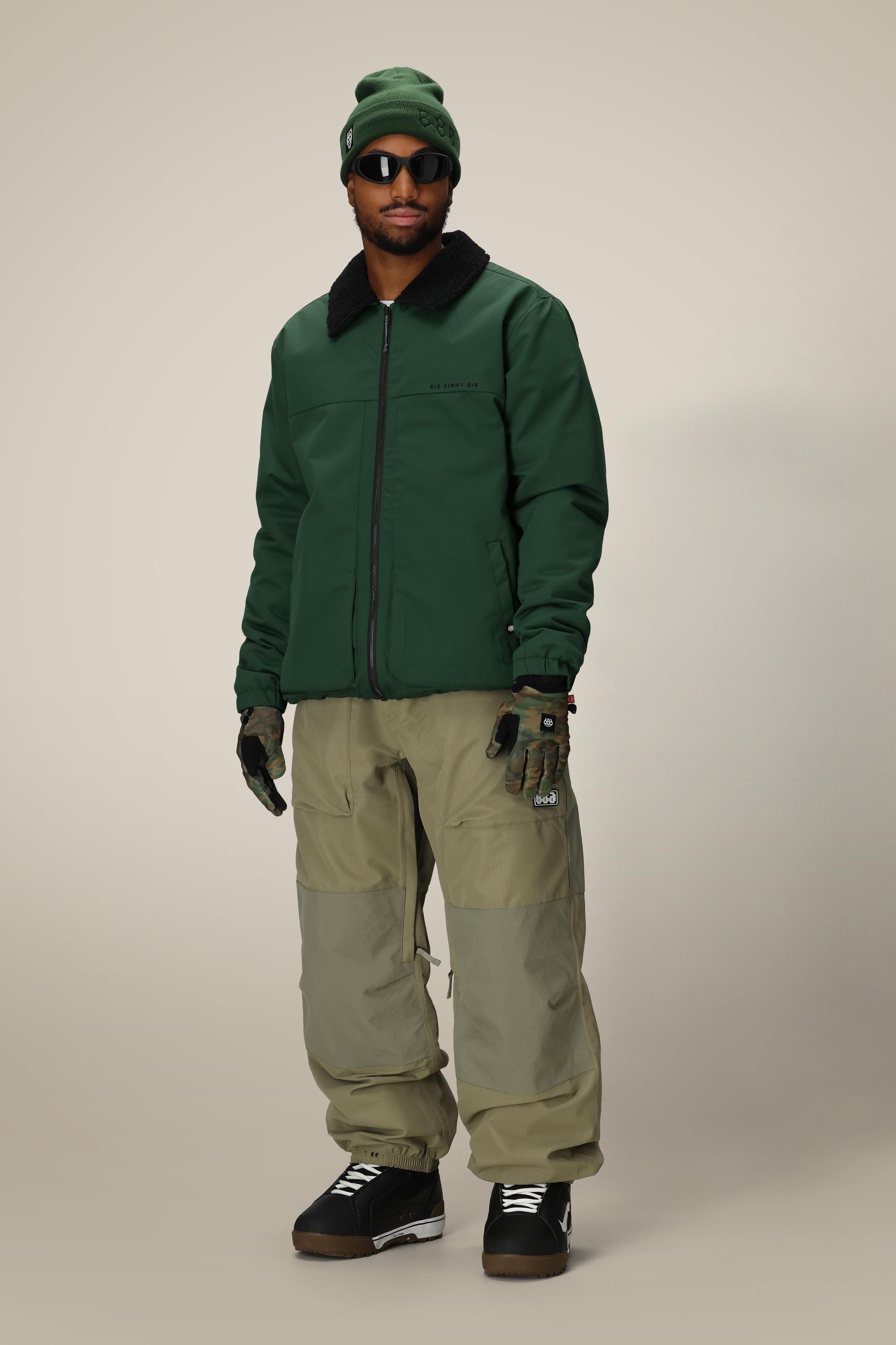 A person wearing the 686 Mens Caim Insulated Jacket stands facing forward against a plain light background, paired with olive green pants, black boots, gloves, a green beanie, and sunglasses.