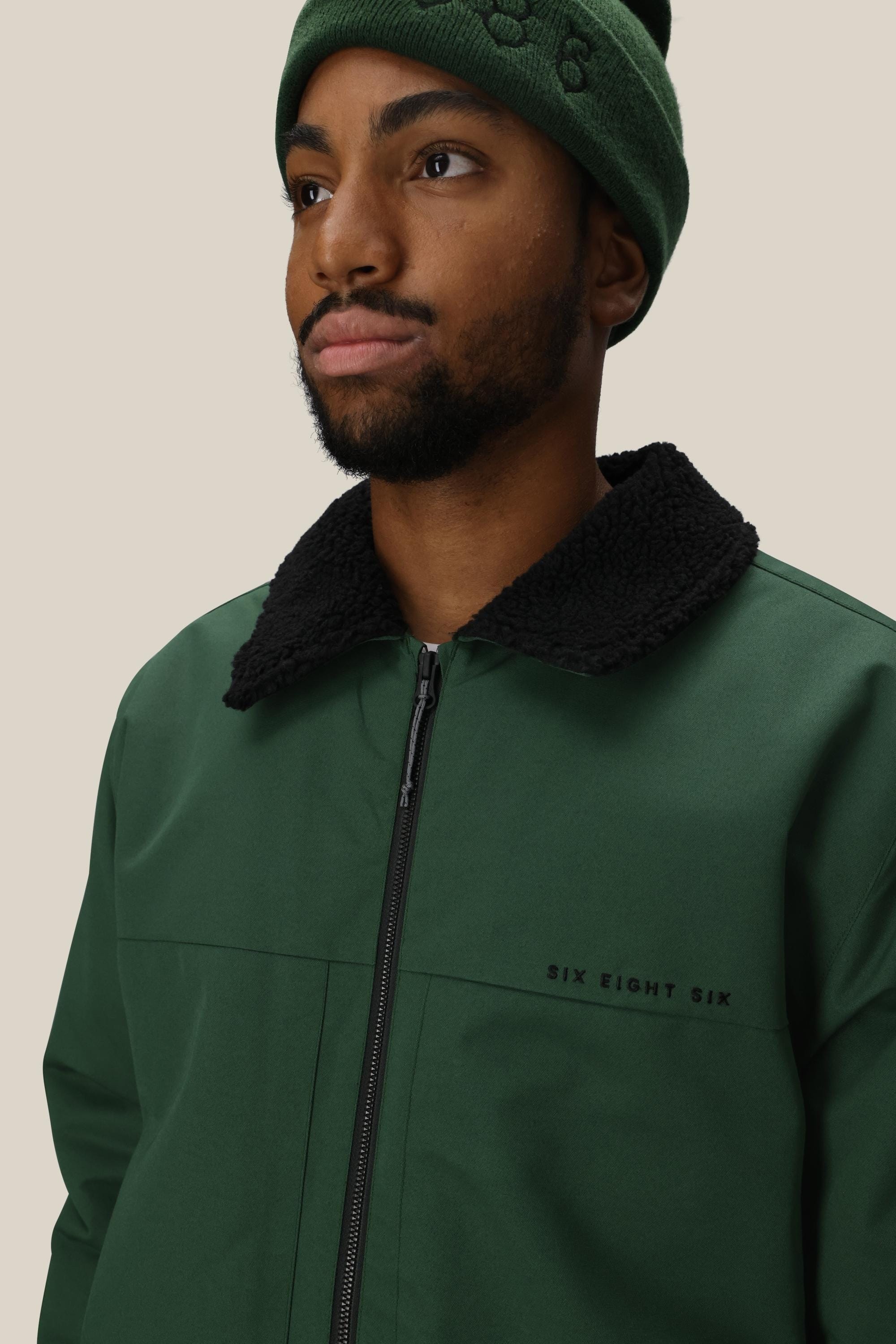 A man in the 686 Mens Caim Insulated Jacket and a green beanie looks slightly to the side. The black fleece collar and SIX EIGHT SIX logo on the chest stand out against a plain, light background.