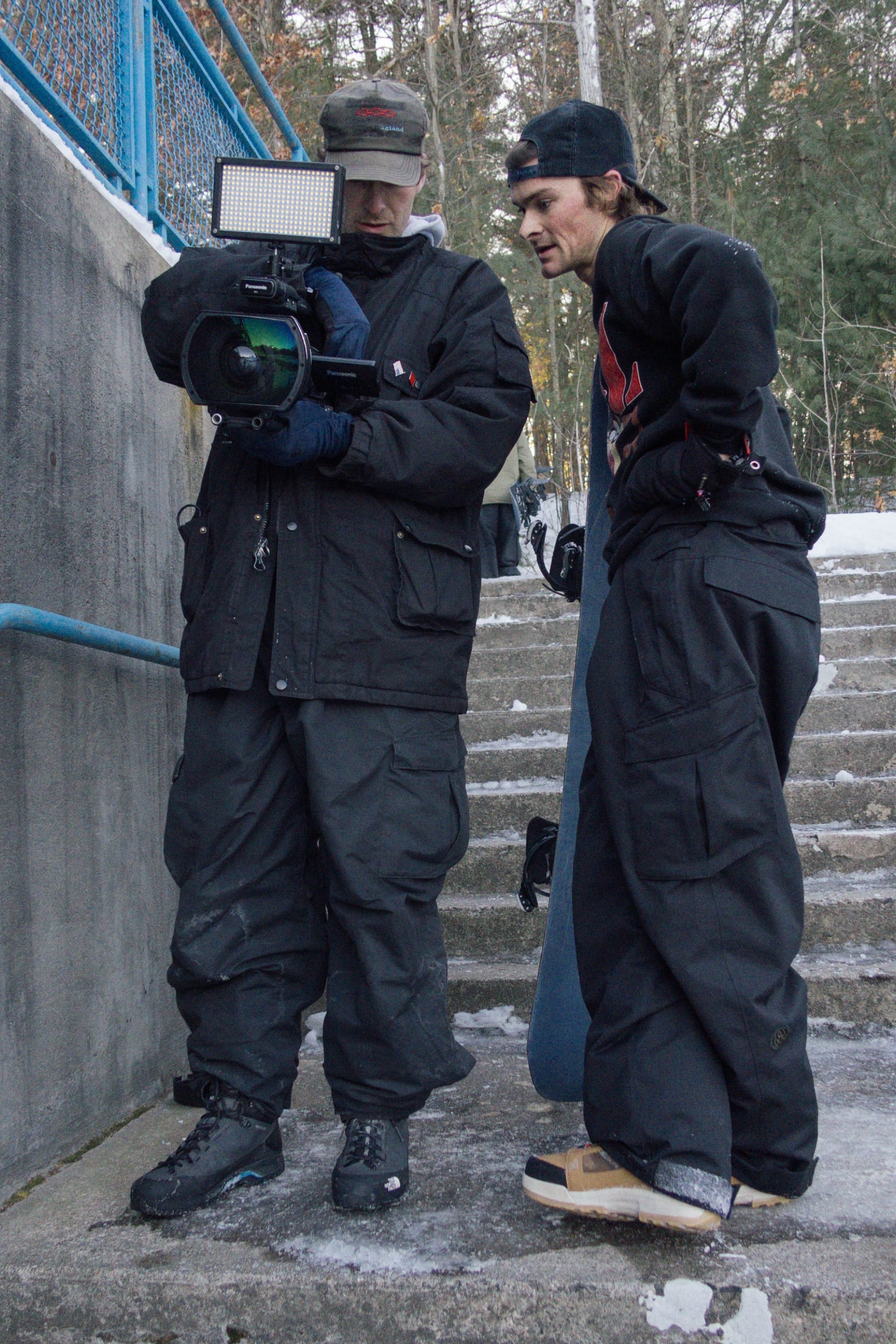 Two people in winter gear stand on snowy outdoor stairs. One, wearing 686 Men's Essox™ Cargo Pant by 686 with cuff adjusters, holds a video camera as the other checks the screen. Trees are visible in the background.