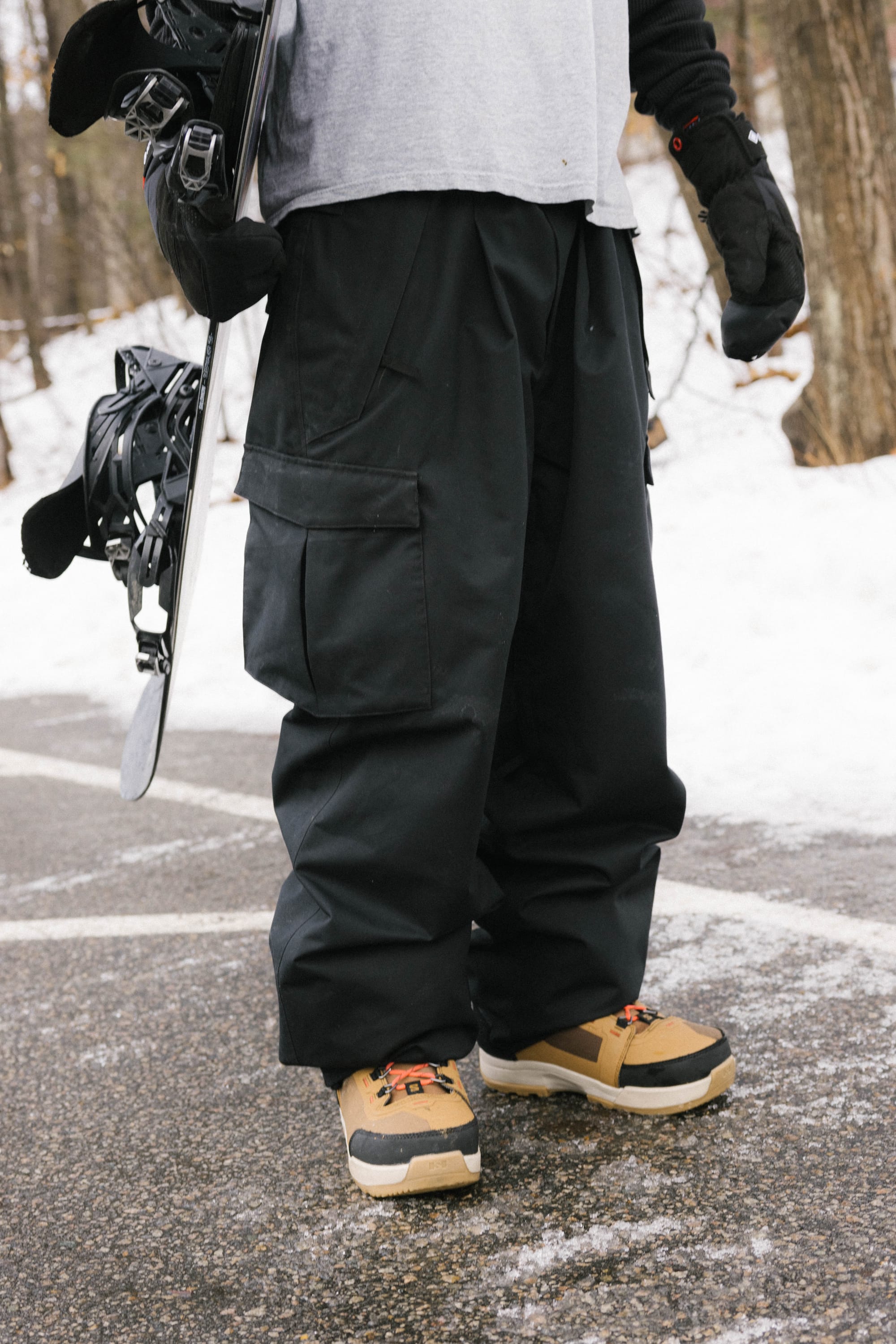 Wearing 686 Men's Essox™ Cargo Pant, tan and black boots, and black gloves, a person holds a snowboard while standing on a snowy path among trees.