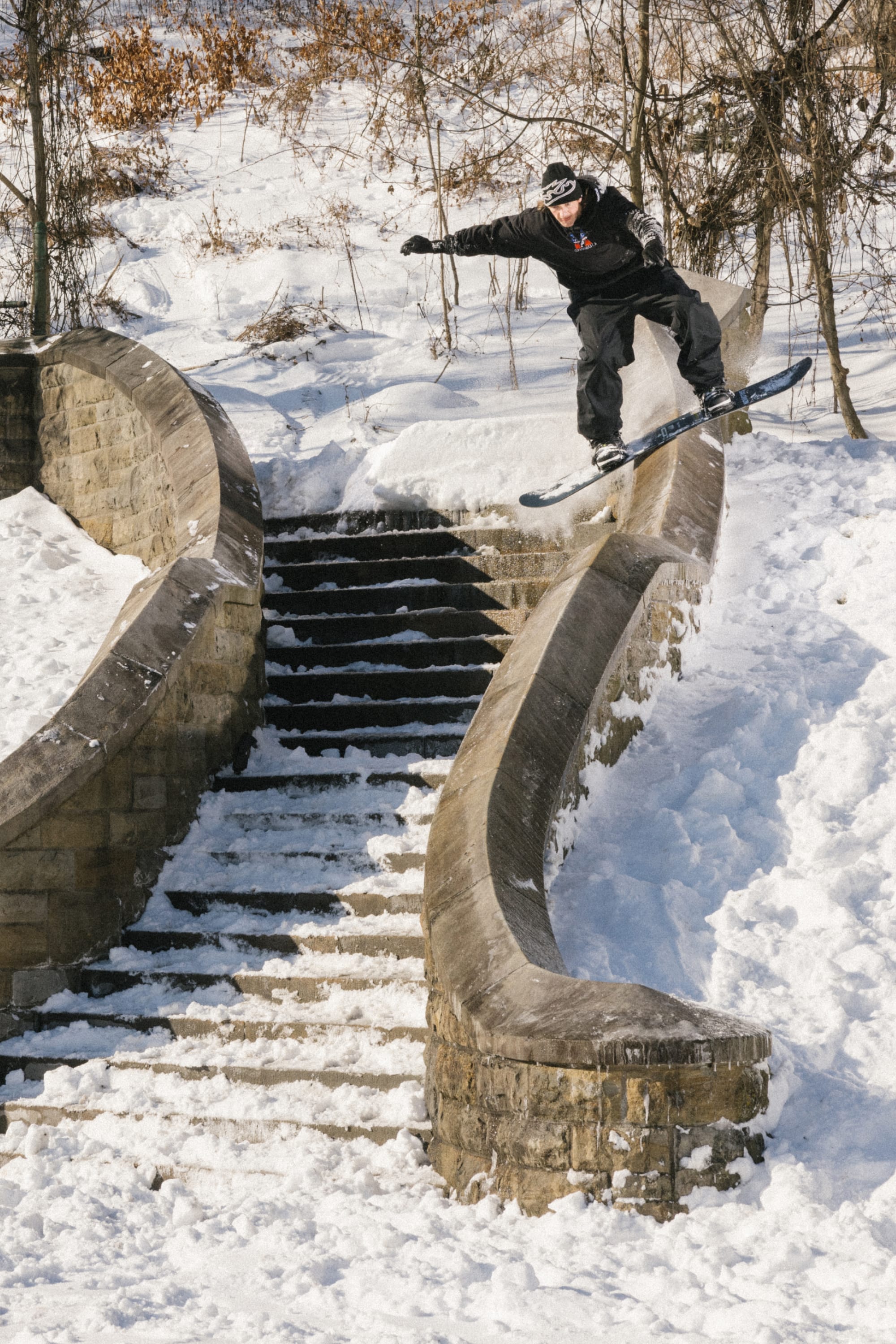 Wearing the 686 Men’s Essox™ Cargo Pant, a snowboarder launches off a snowy stone stair railing, balancing midair with arms outstretched as fresh snow blankets the steps and surrounding landscape.