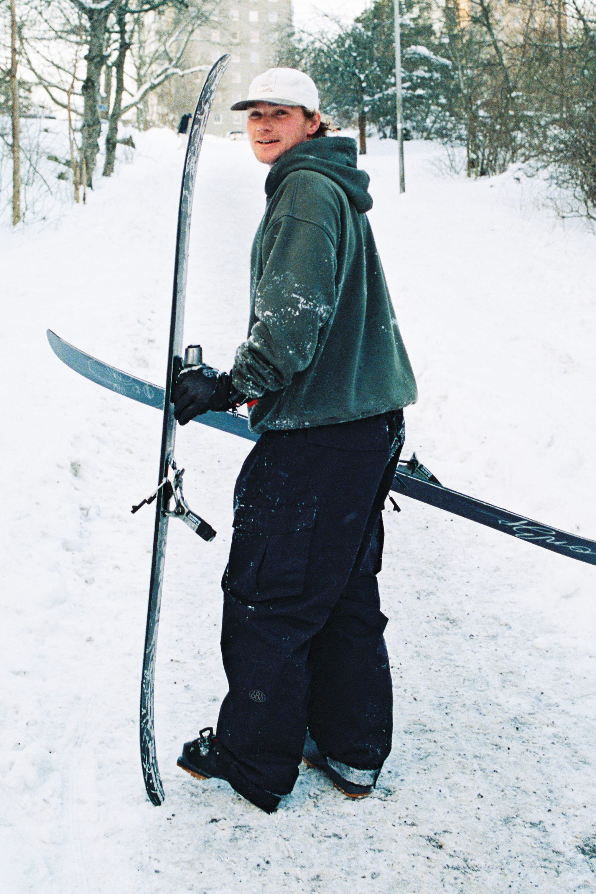 Wearing the 686 Men’s Essox™ Cargo Pant, a person in winter clothing and a white cap smiles while holding a pair of skis on a snowy path surrounded by trees.