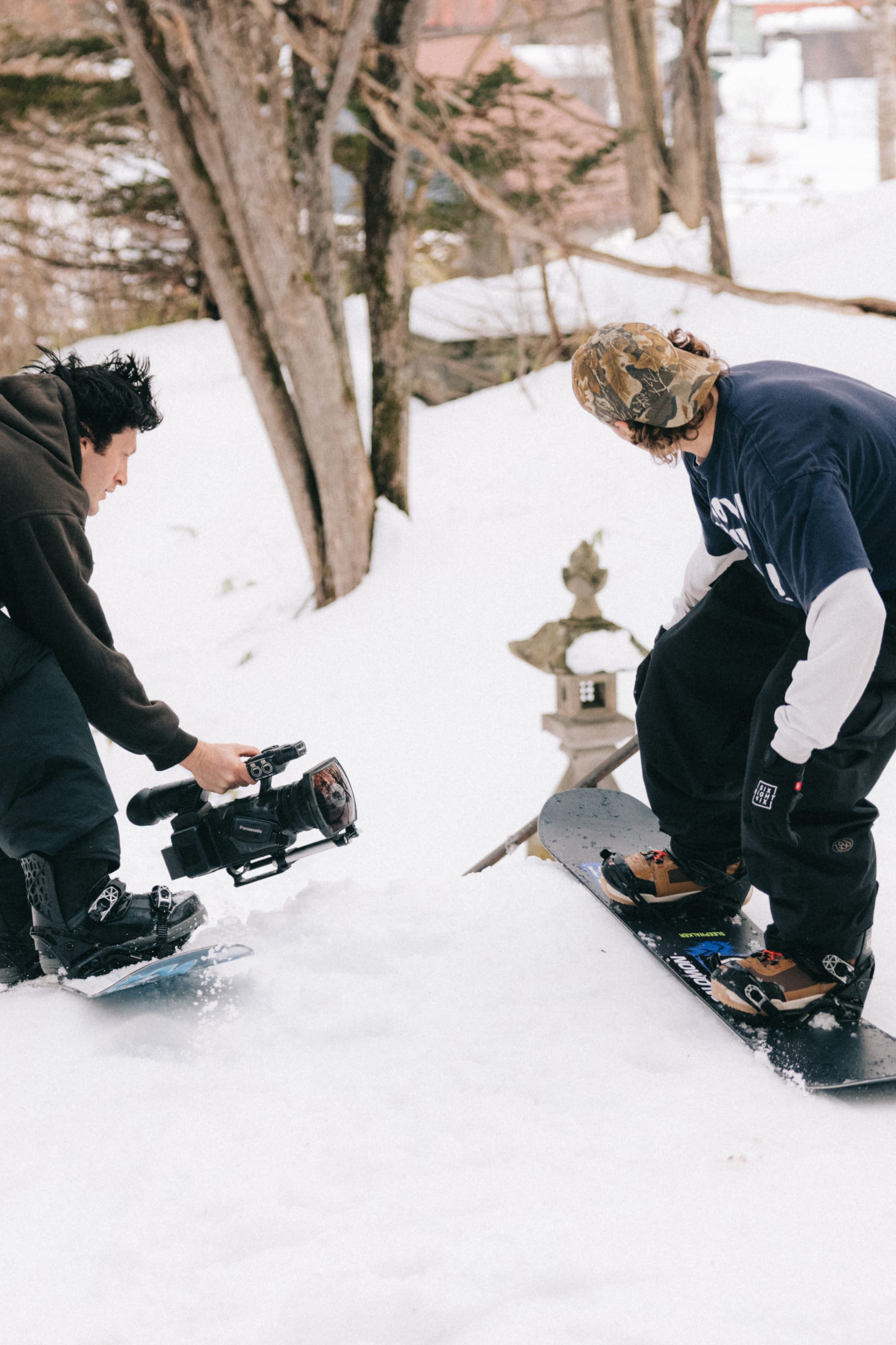 A person with a camera films a snowboarder in winter clothing and 686 Men's Essox™ Cargo Pant by 686, standing on a snowboard in the snow with trees and a small snow-covered structure in the background.