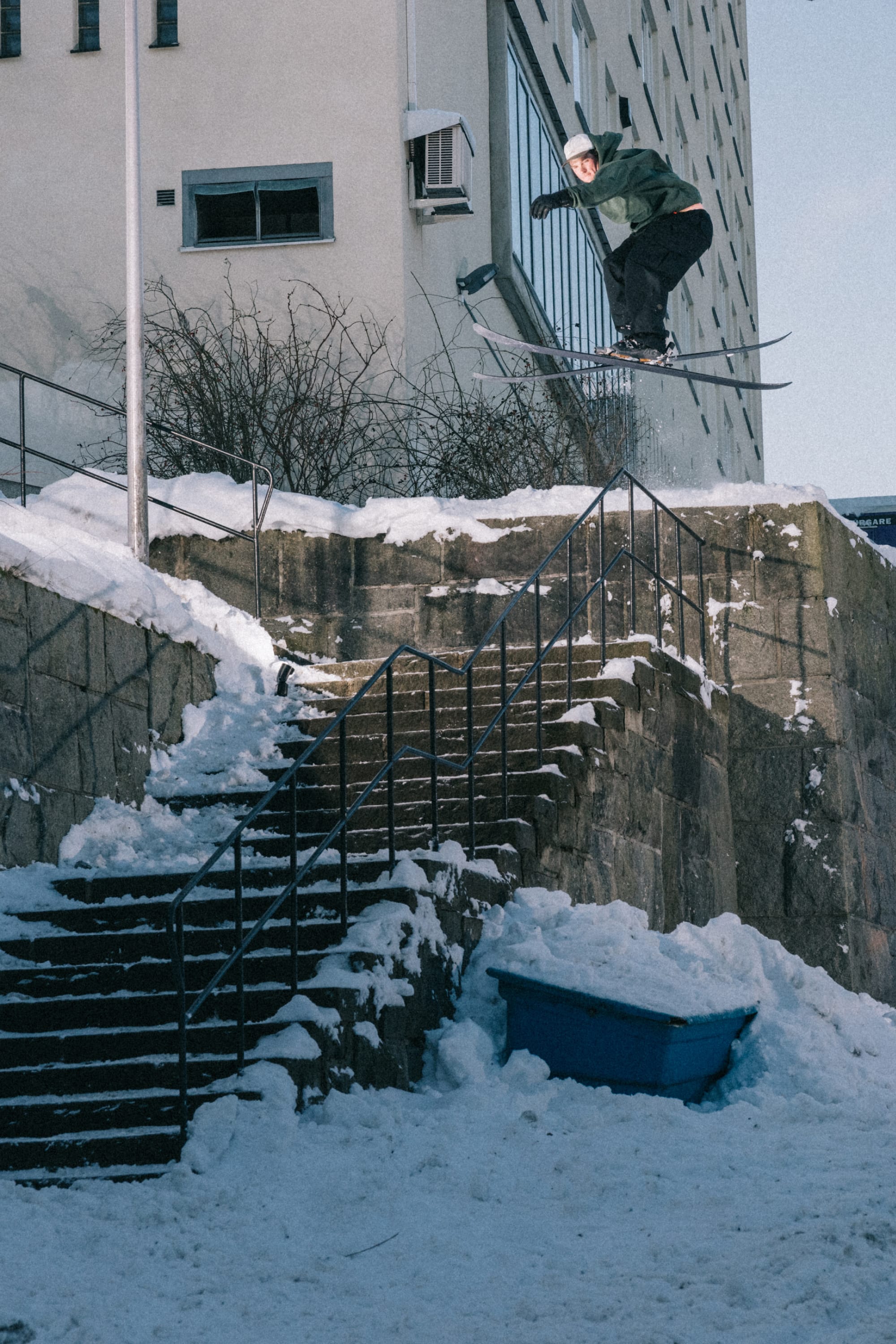 A skier wearing a green jacket, white helmet, and 686 Men’s Essox™ Cargo Pant launches over a snowy stone stairway beside a city building on a winter day.