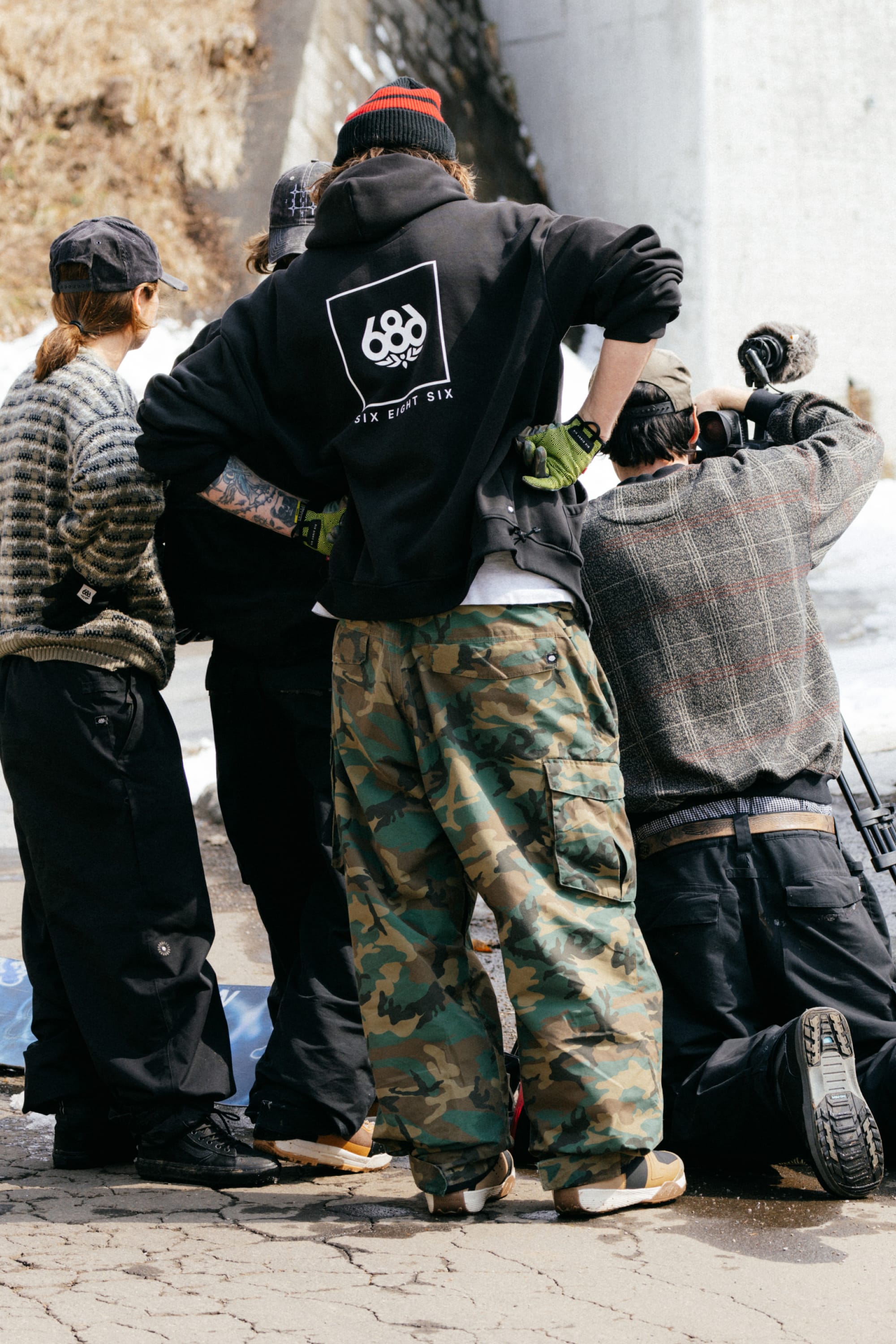 Four people in winter clothing stand together outdoors. One wears a black hoodie with a 686 logo and the 686 Men's Essox™ Cargo Pant, featuring adjustable cuffs and tapered legs.