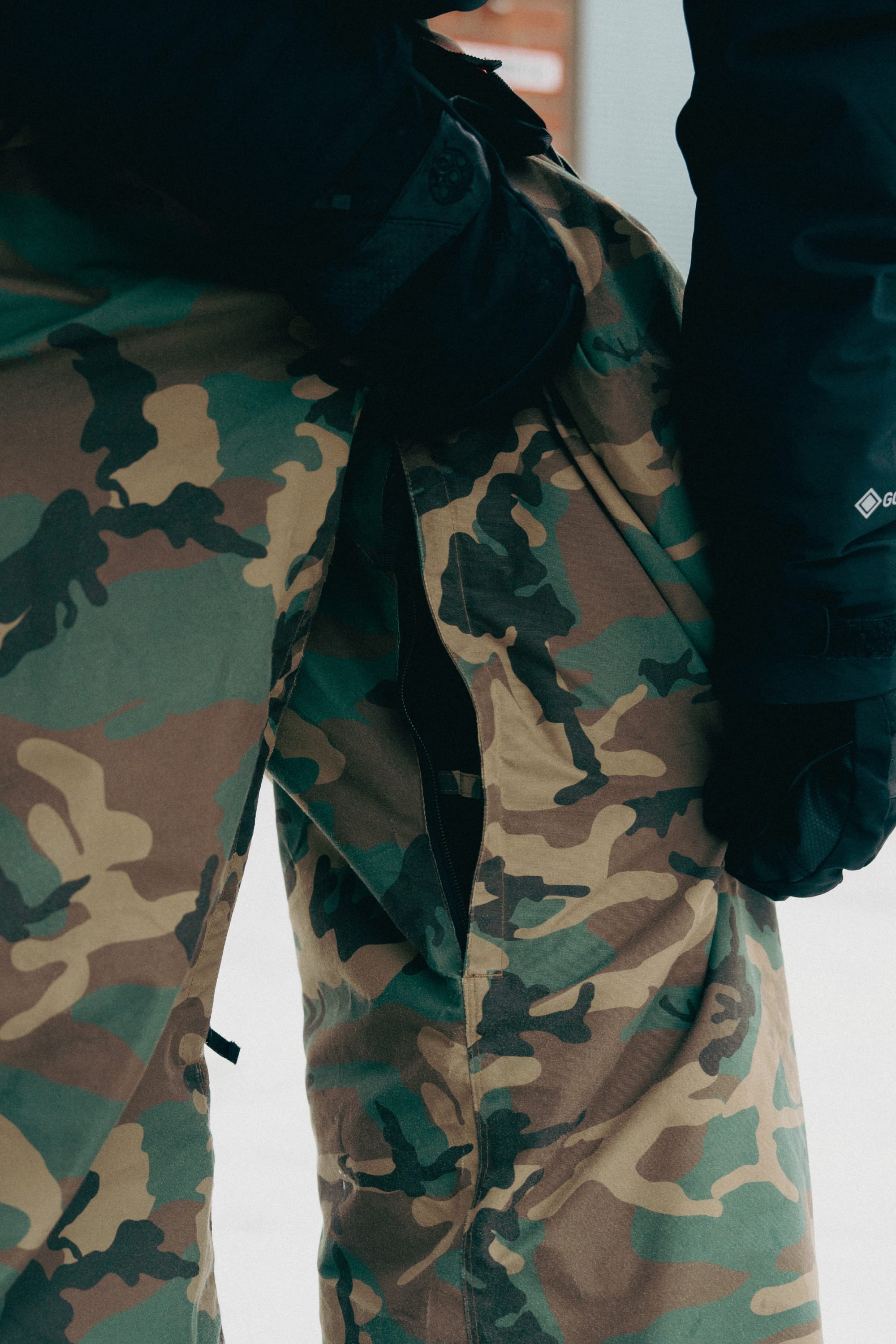 A person wearing the 686 Men’s Essox™ Cargo Pant by 686, featuring a large tear along the inner thigh that reveals black fabric underneath. They are also wearing black gloves and a dark jacket.