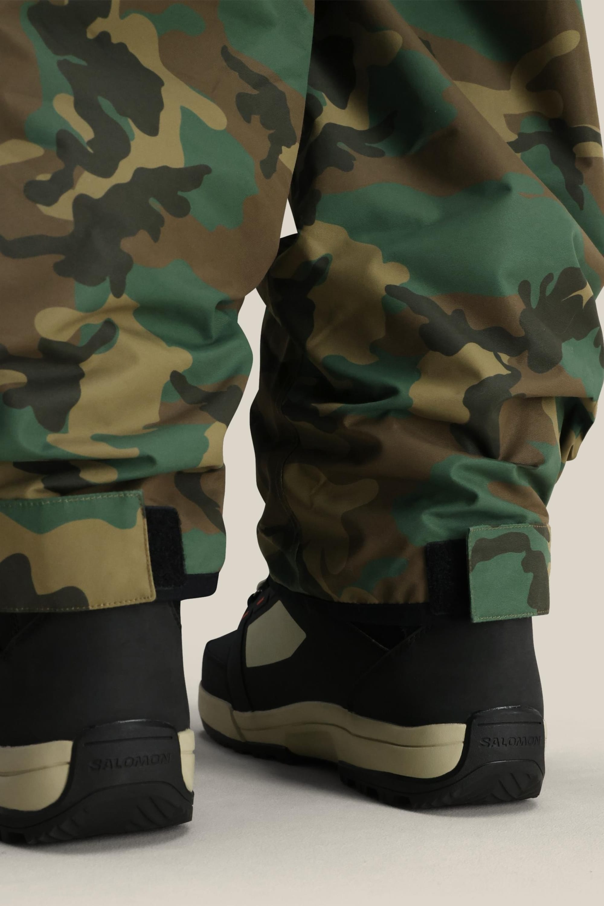 Close-up of a person wearing 686 Men’s Essox™ Cargo Pant in camouflage and black boots, standing on a light surface. The photo is focused on the lower legs and feet.