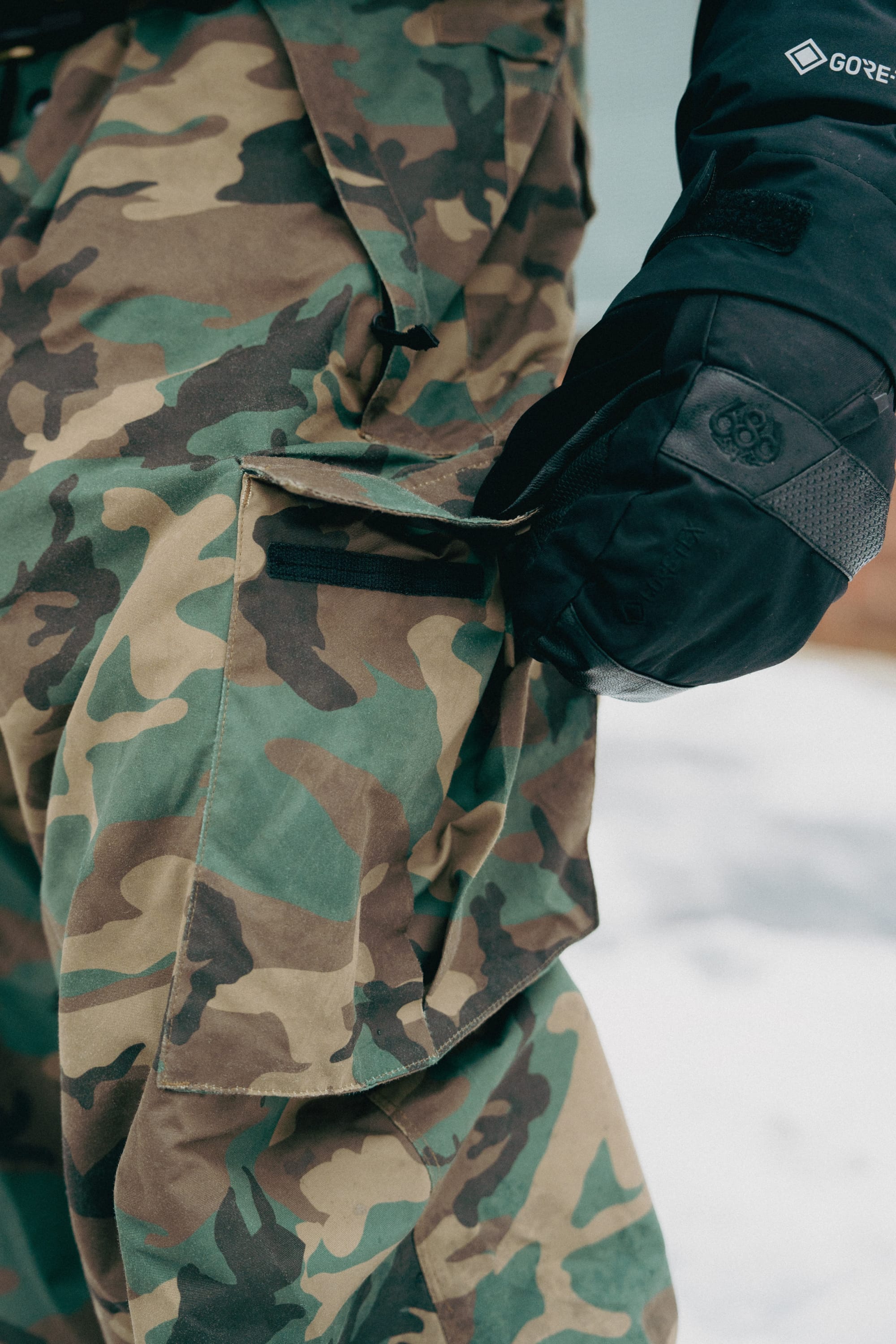 A person wearing 686 Men's Essox™ Cargo Pant with tapered legs and a black glove opens a cargo pocket flap, with snow visible on the ground in the background.