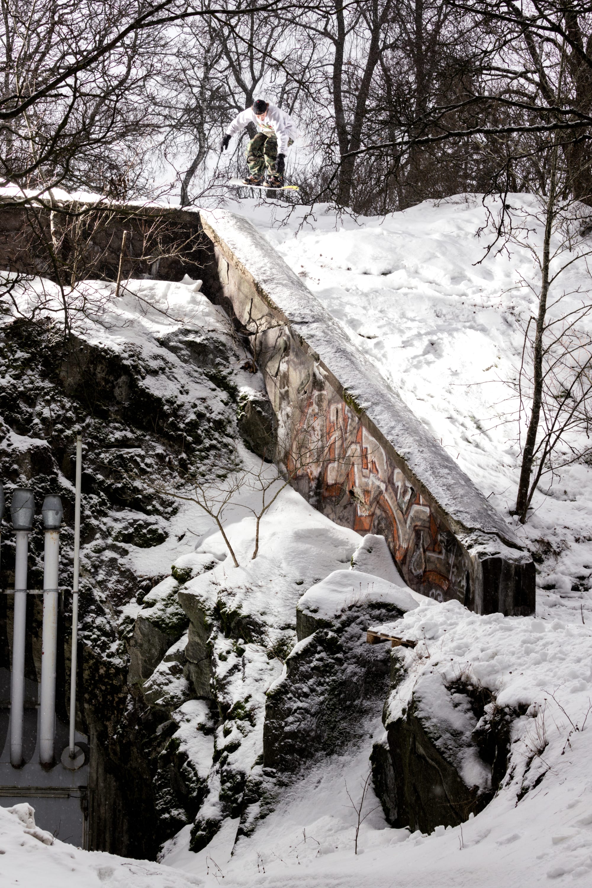 Wearing the 686 Men’s Essox™ Cargo Pant, a skier launches off a snow-covered, graffiti-marked concrete ledge in a winter landscape framed by bare trees and rocks.