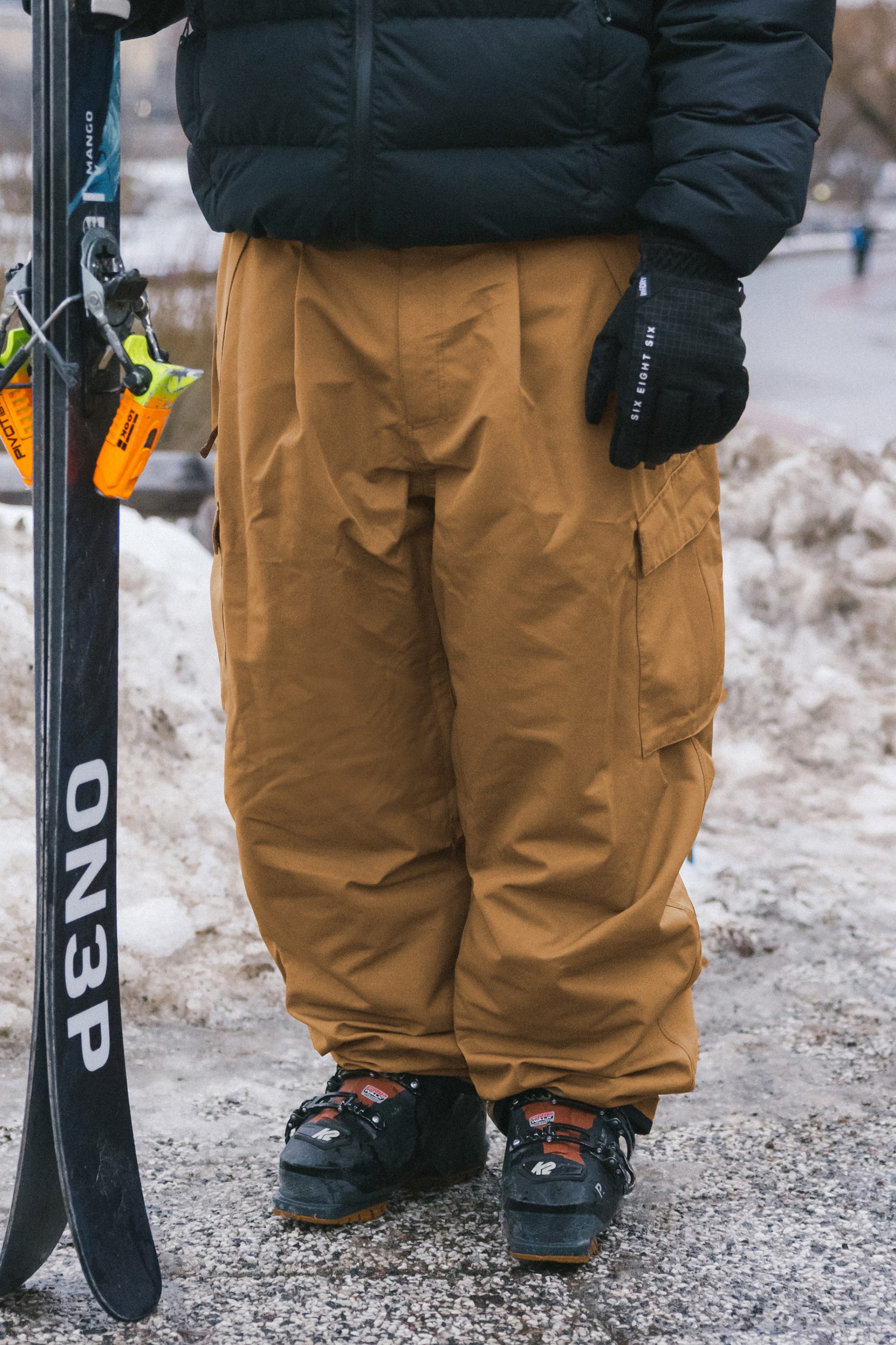 A person wearing 686 Men’s Essox™ Cargo Pant in tan, paired with a black jacket and gloves, holds ON3P skis while standing on snow in ski boots.
