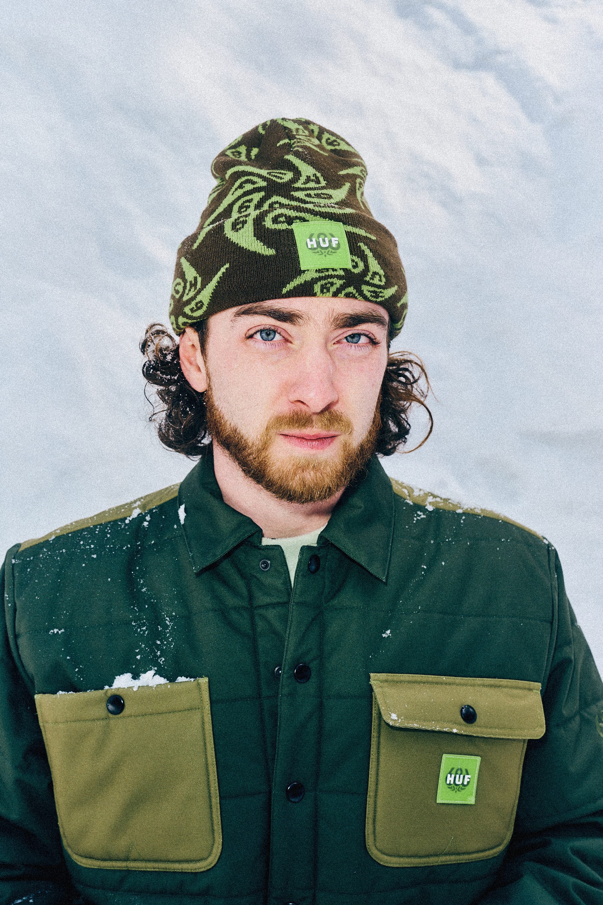 A man with curly hair and a beard stands outside in the snow, wearing a patterned green 686 HUF Beanie and a mid-weight dark green jacket with olive pockets, both featuring branding.