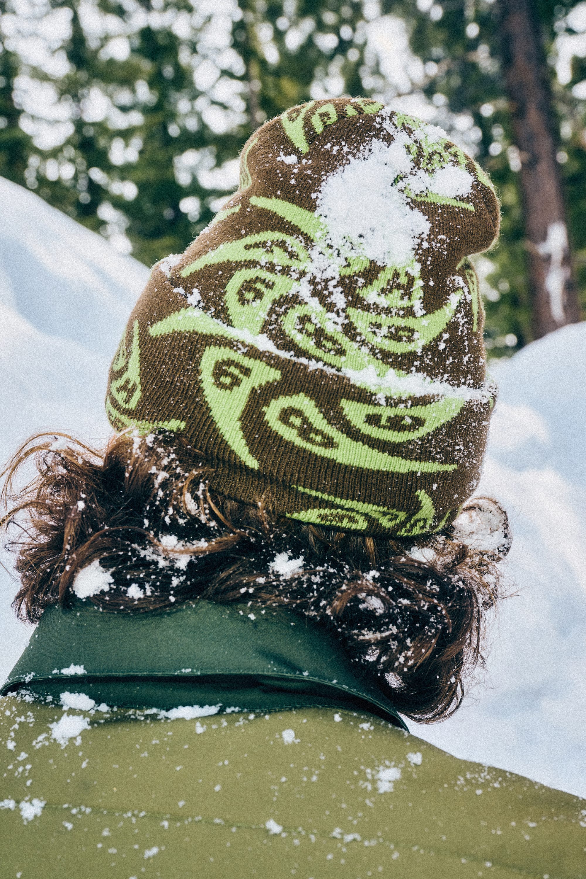 A person with curly hair stands outdoors in a green patterned 686 HUF Beanie by 686 and an olive jacket, snow covering their hat and shoulders. Snow-laden trees appear behind them as they face away from the camera.