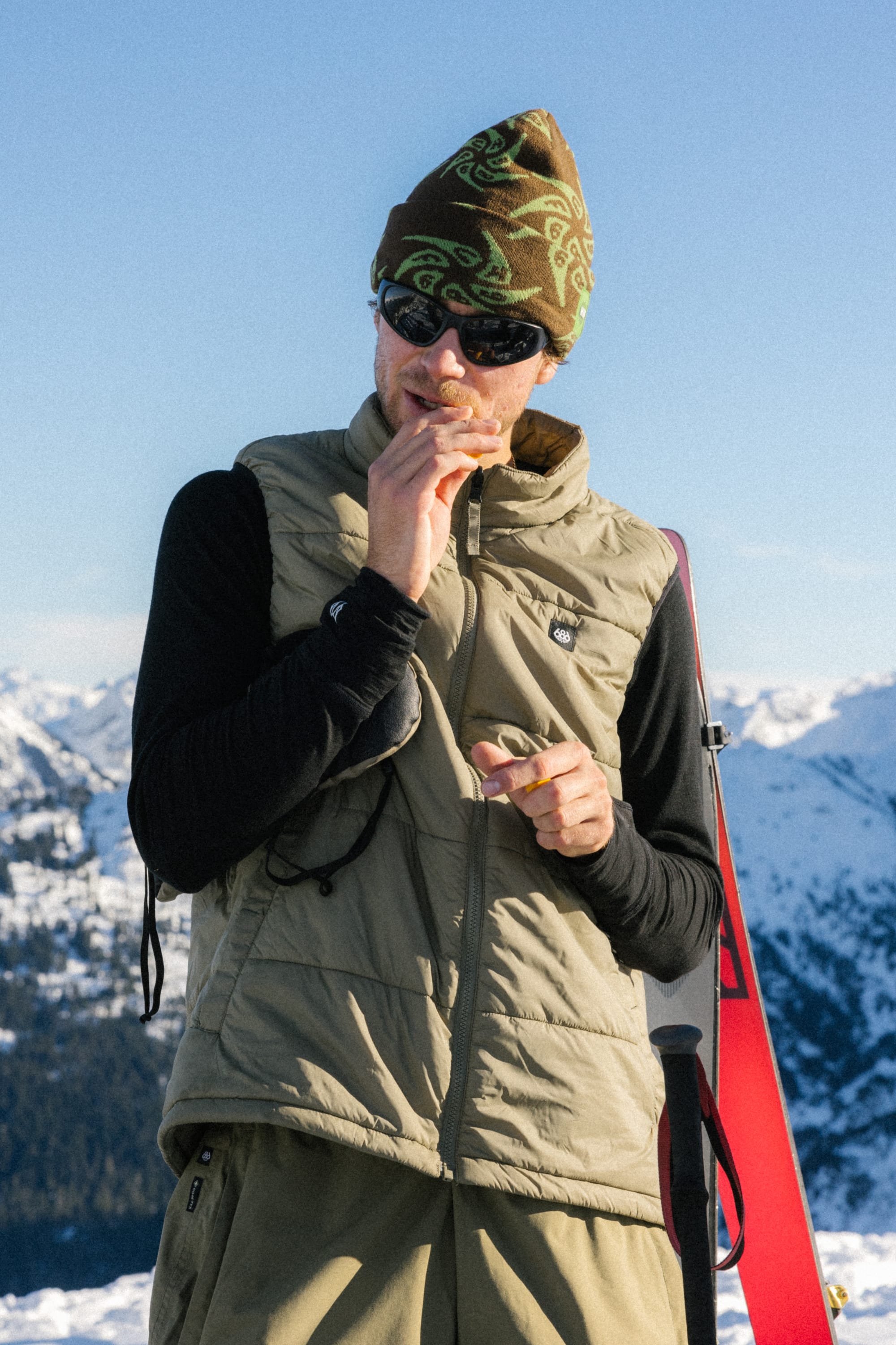 Wearing a 686 HUF Beanie and ski gear, a person enjoys a snack on a snowy mountain, surrounded by snow-covered peaks under a clear blue sky.