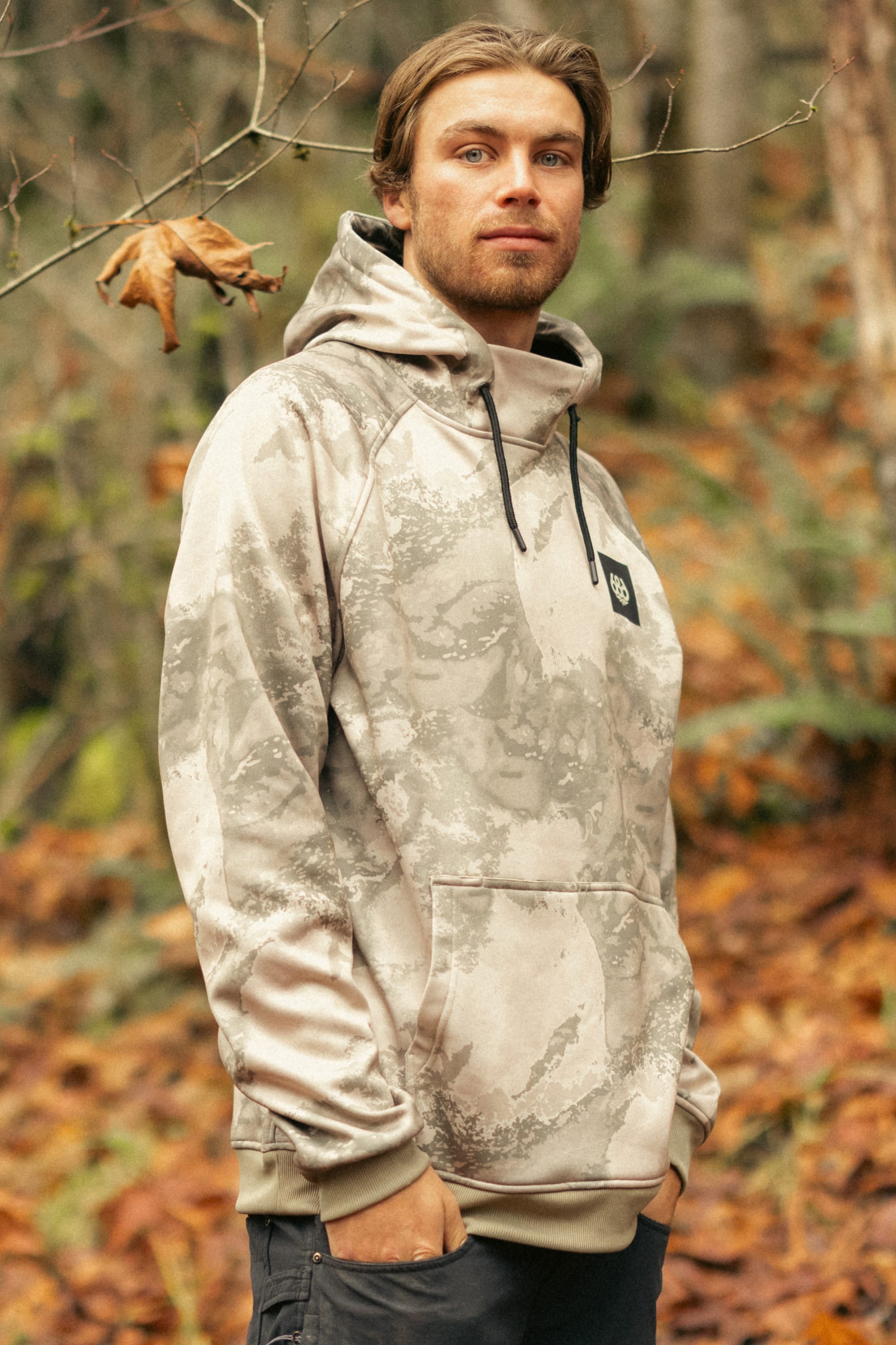 A man with light brown hair and a beard stands in a forest wearing the 686 Mens Bonded Fleece Pullover Hoody. This light gray and beige camo hoodie by 686 features fleece lining, ideal for cool autumn days outdoors.