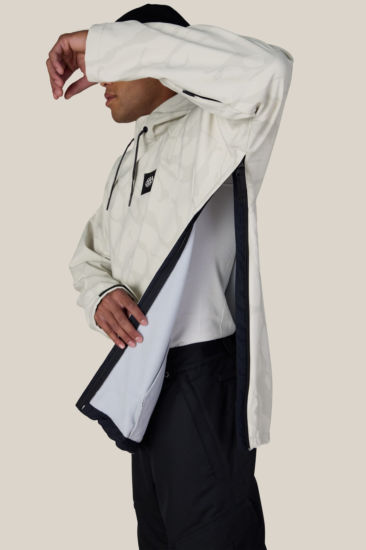A man models the 686 Men’s Waterproof Hyperchromic Pullover Hoody in a light color with black pants, lifting one arm to reveal the large side zipper opening and the inner lining of the hoody against a plain background.