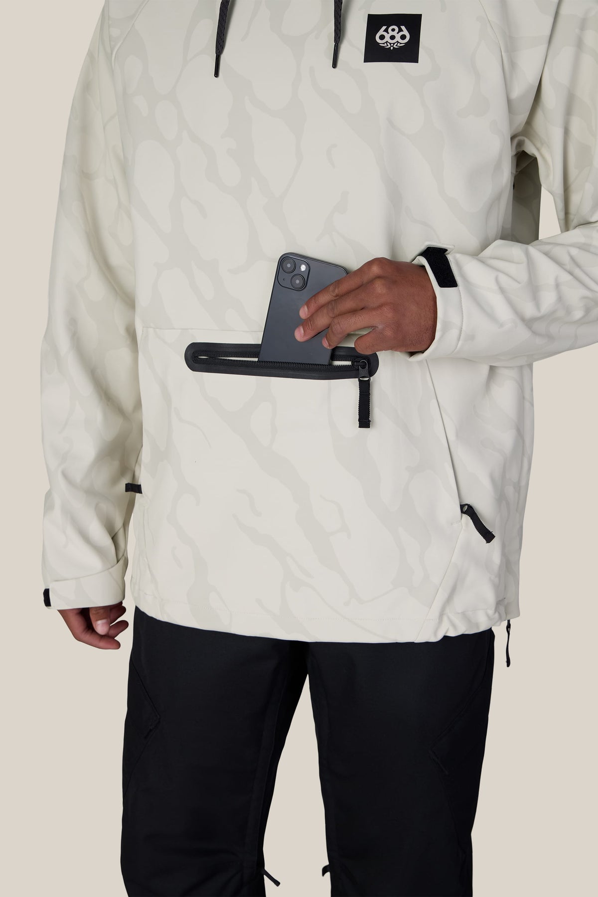 A person wearing the 686 Men’s Waterproof Hyperchromic Pullover Hoody and black pants puts a smartphone into a horizontal zippered pocket on the front of the hoodie.