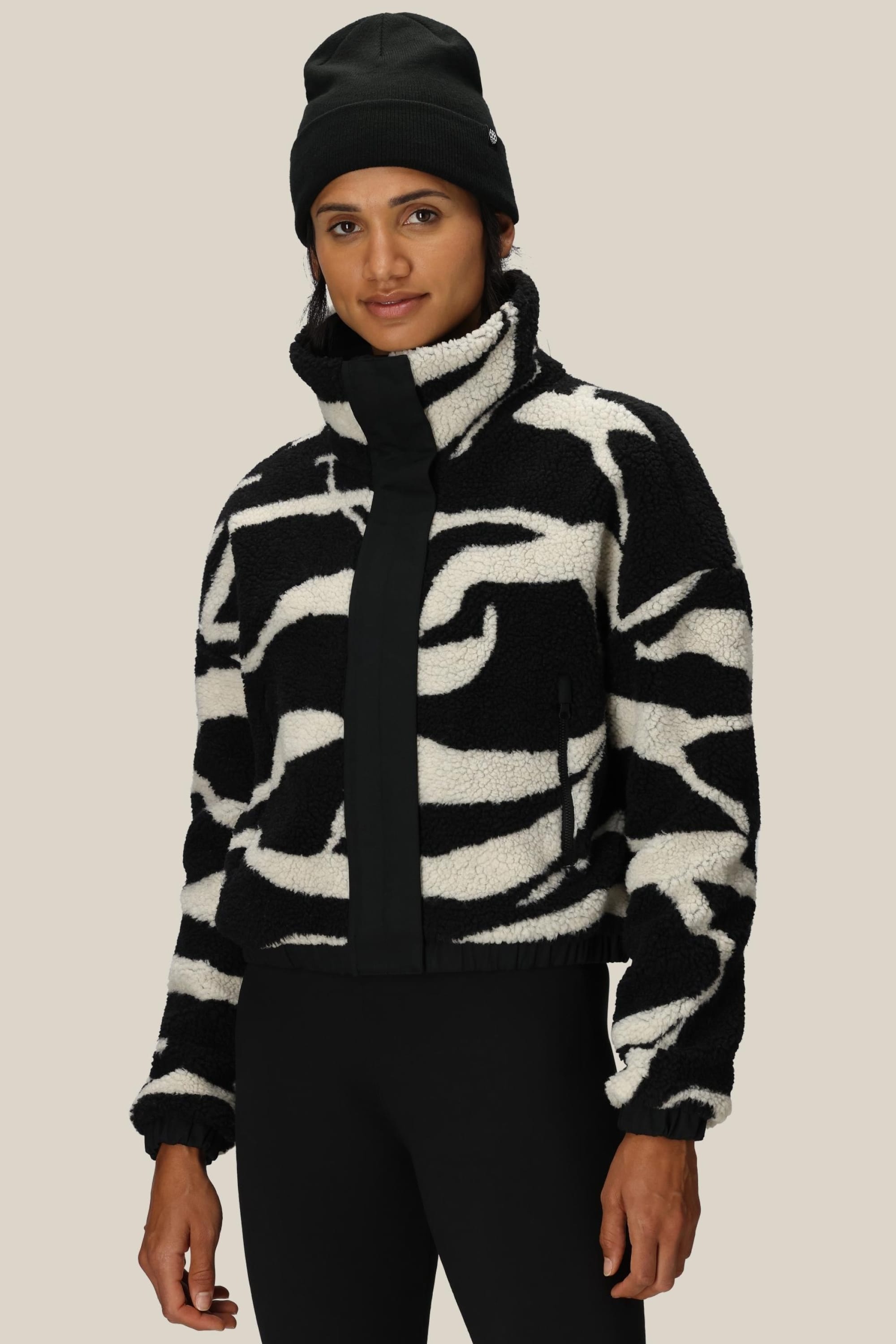 A person wears a black beanie, black pants, and the 686 Womens Outpost Sherpa Jacket by 686—a bold black-and-white high-collar fleece with swirl patterns—while standing against a plain beige background.