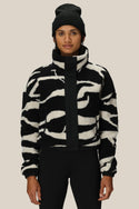 A person wearing a black beanie and the 686 Womens Outpost Sherpa Jacket by 686, featuring a cozy black-and-white pattern, stands facing forward against a neutral background.