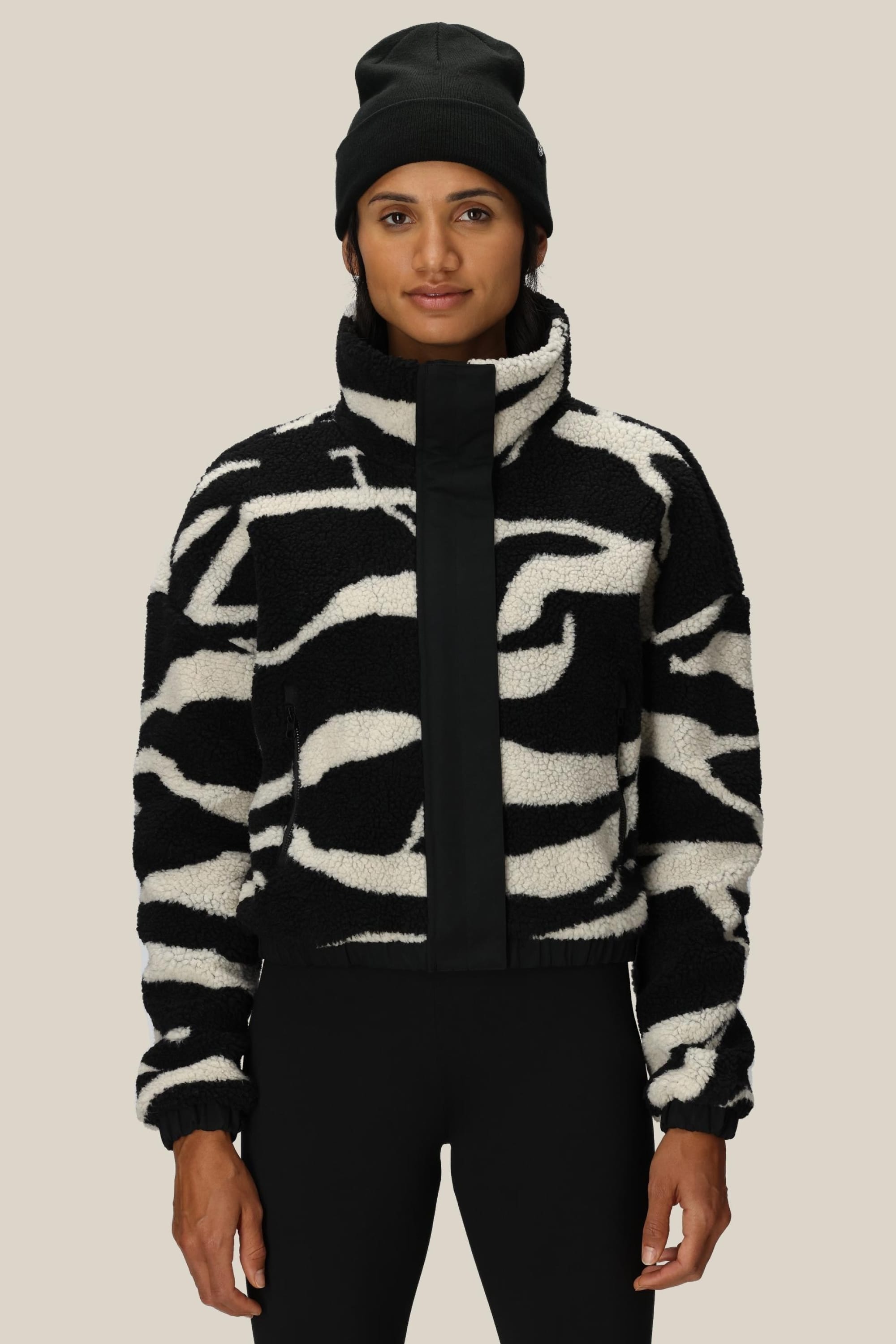 A person wearing a black beanie and the 686 Womens Outpost Sherpa Jacket by 686, featuring a cozy black-and-white pattern, stands facing forward against a neutral background.