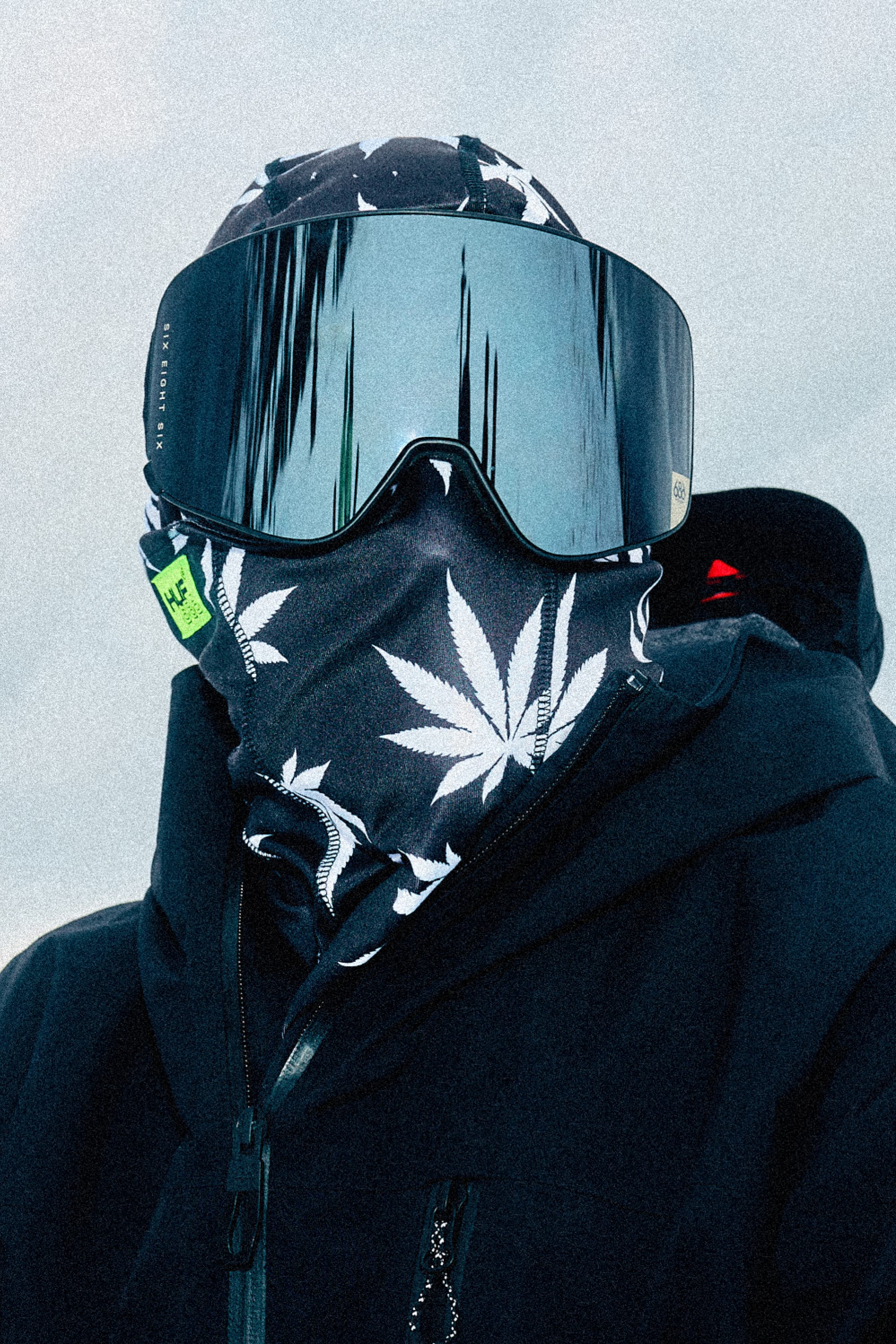 A person in a black jacket and large reflective ski goggles wears the 686 Alpha Hinged Balaclava by 686, featuring white cannabis leaf patterns, while standing outside in the snow.