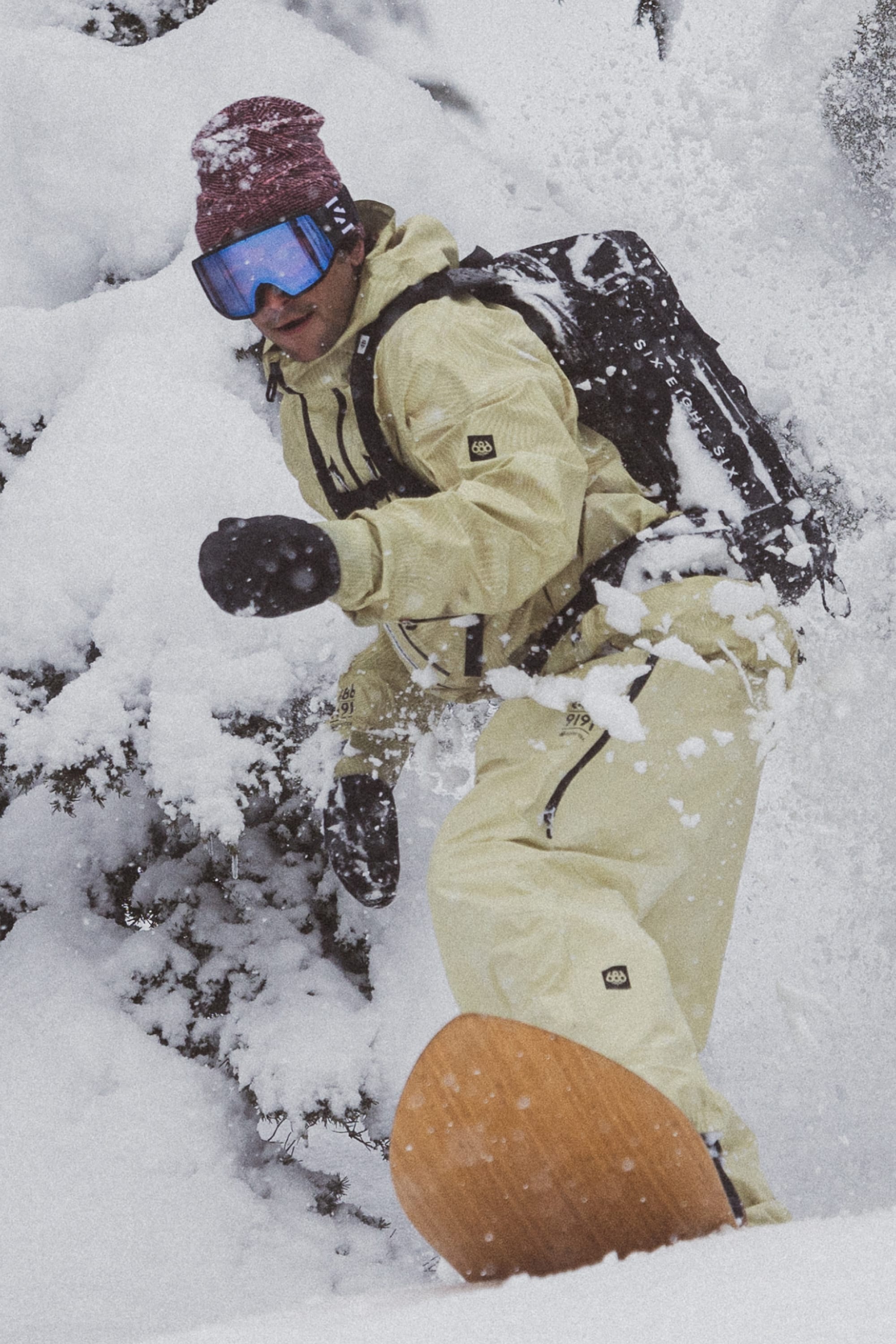 A snowboarder in beige outerwear, a maroon beanie, blue goggles, and 686 Men's GORE-TEX Leather Linear™ Mitts rides through fresh snow on a mountainside amid snow-covered trees.