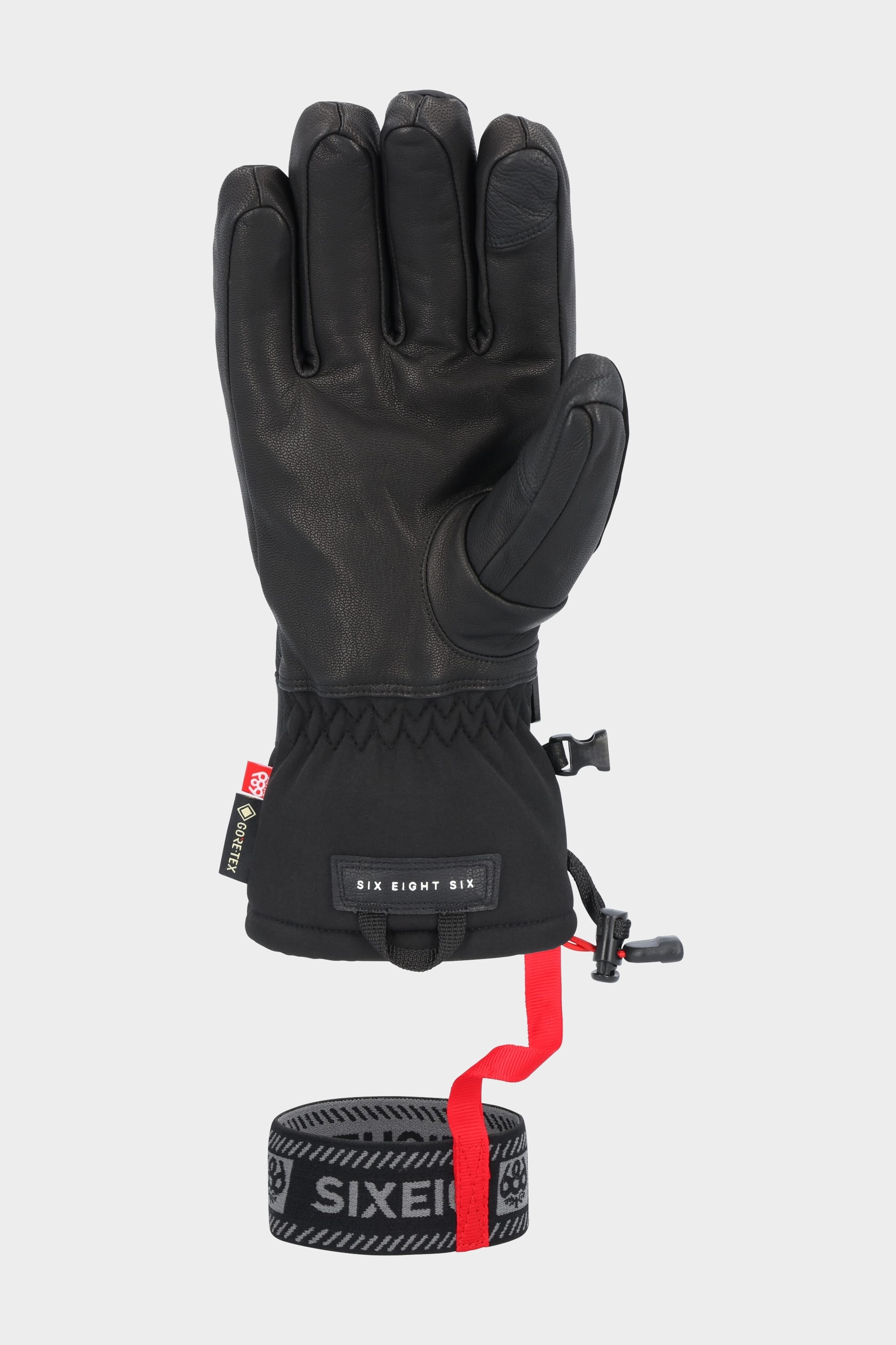 The 686 Men's GORE-TEX Leather Linear™ Glove in black features leather palm detailing, an adjustable wrist strap, and a removable black and gray wrist leash with the 686 brand name and a red connecting strap. The glove is shown upright on a plain background.