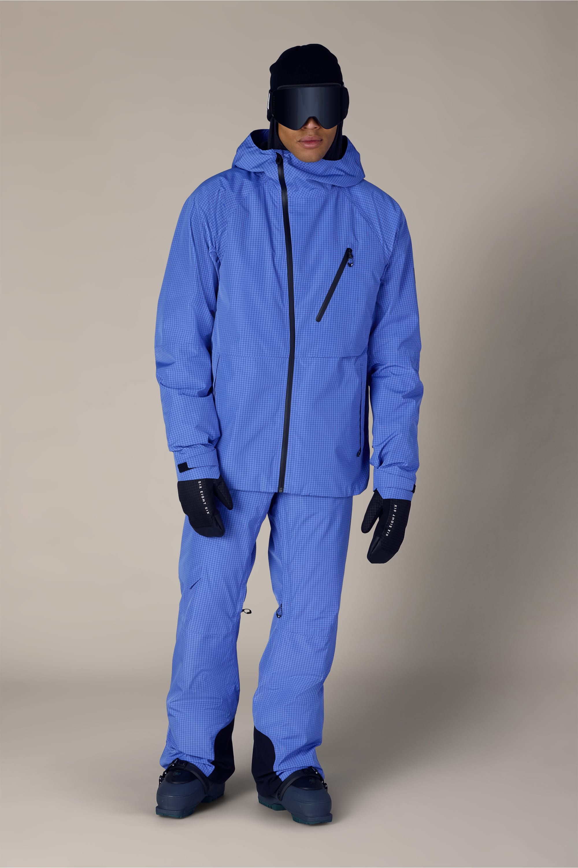 A person wearing the 686 Men’s Hydra™ Thermagraph® Jacket in blue, along with matching pants, black gloves, boots, and a black helmet with large black ski goggles, standing against a beige background.
