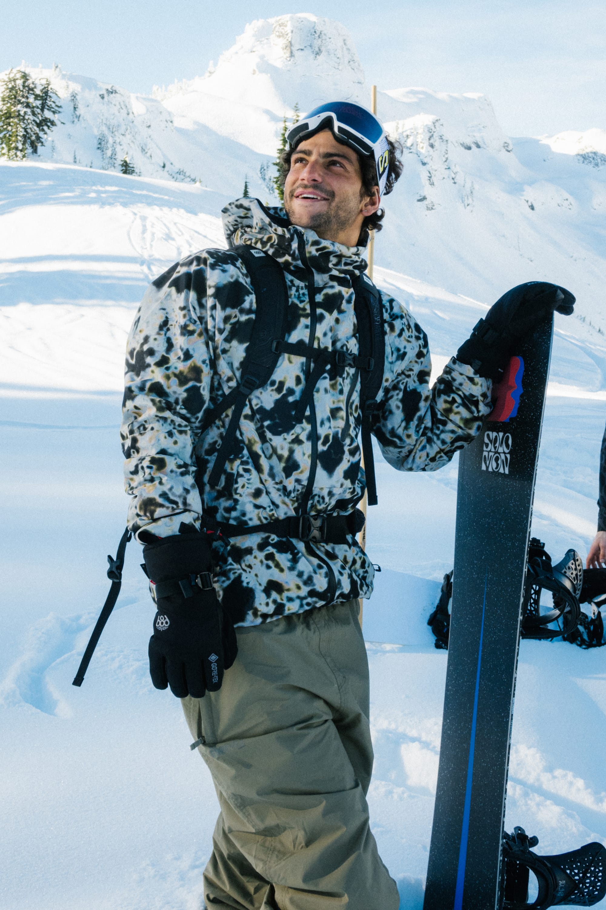 A person wearing the 686 Men's Hydra™ Thermagraph® Jacket and goggles smiles while holding a snowboard upright in snowy mountains under a blue sky.