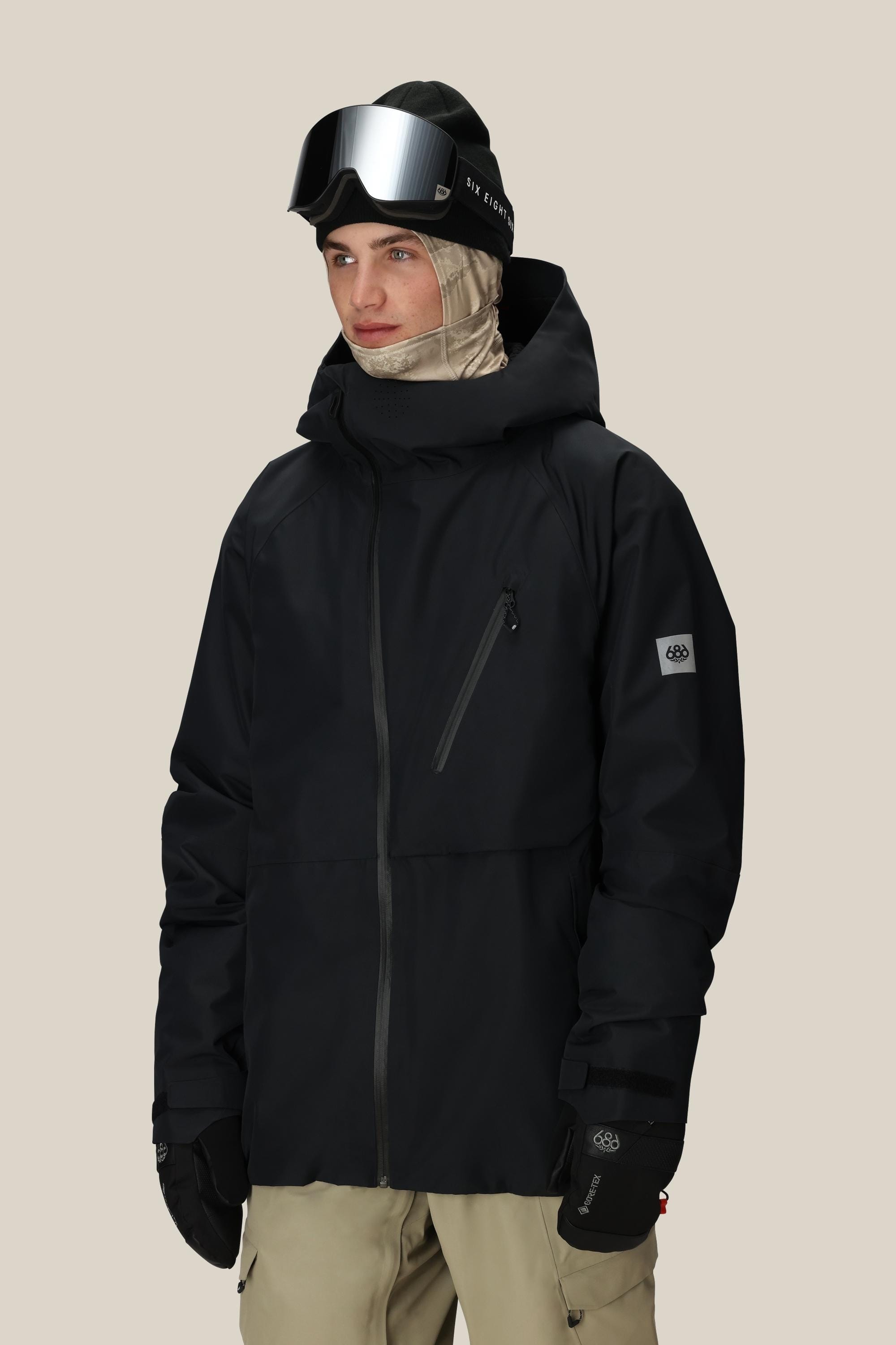 A person wearing the 686 Men’s Hydra™ Thermagraph® Jacket by 686, along with beige snow pants, black gloves, a beige balaclava, and black ski goggles against a plain light background.