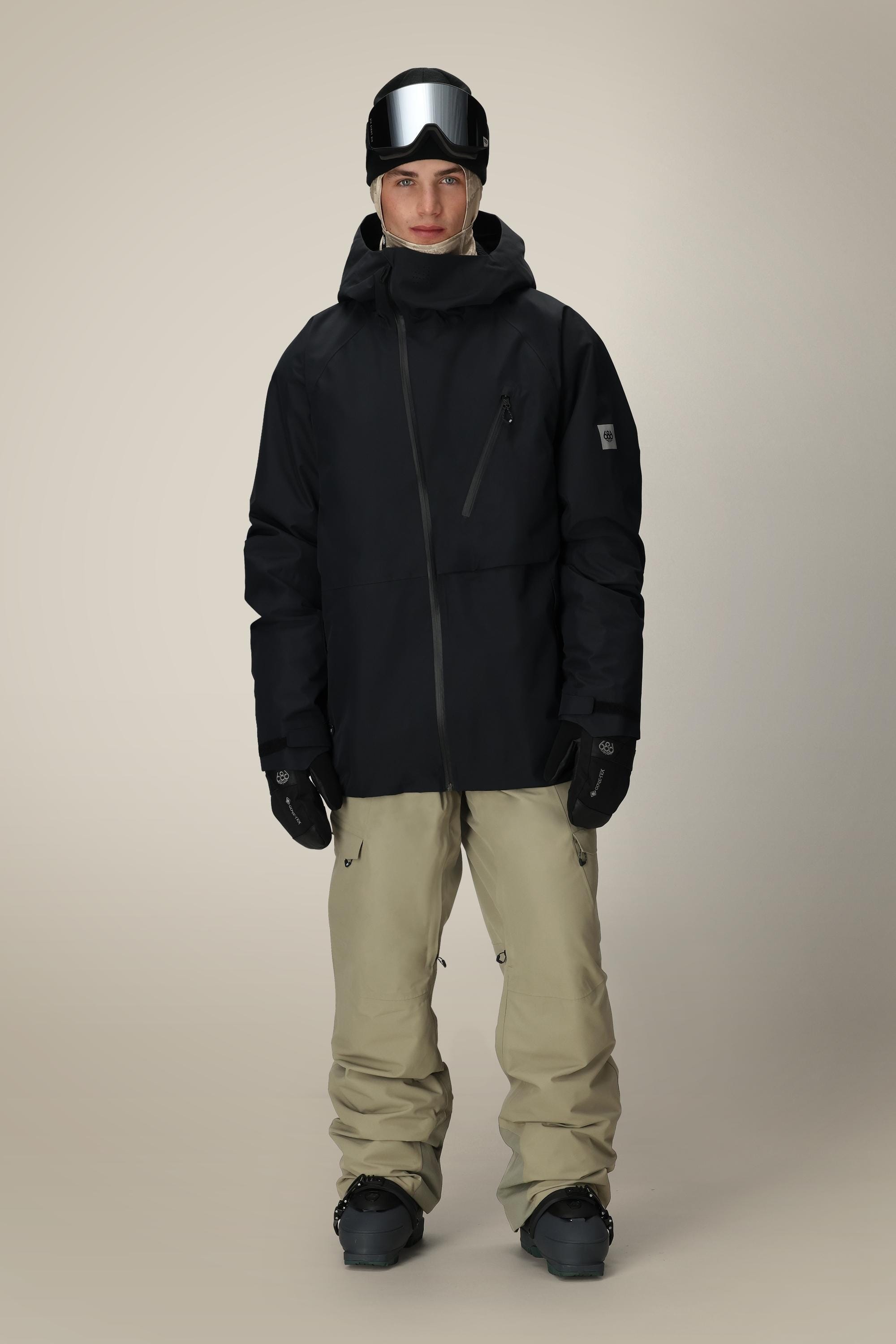 A person wears the 686 Men's Hydra™ Thermagraph® Jacket in black, paired with beige snow pants, black gloves, and a black helmet with reflective ski goggles, standing against a plain light background.