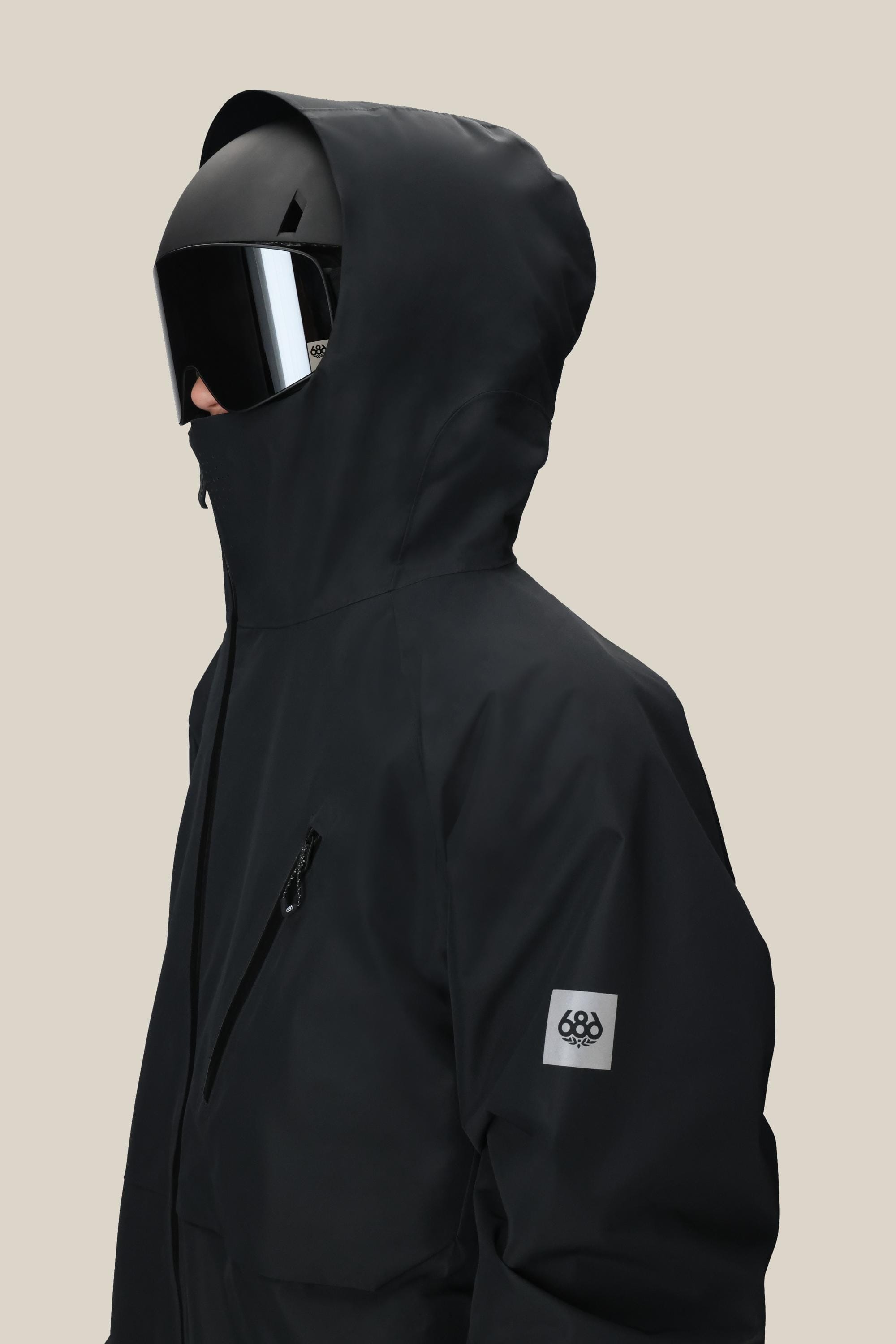 A person wearing the 686 Men’s Hydra™ Thermagraph® Jacket—a black hooded jacket with a high collar, zippered chest pocket, and a small patch logo on the upper sleeve—along with a black helmet and reflective visor, facing left.