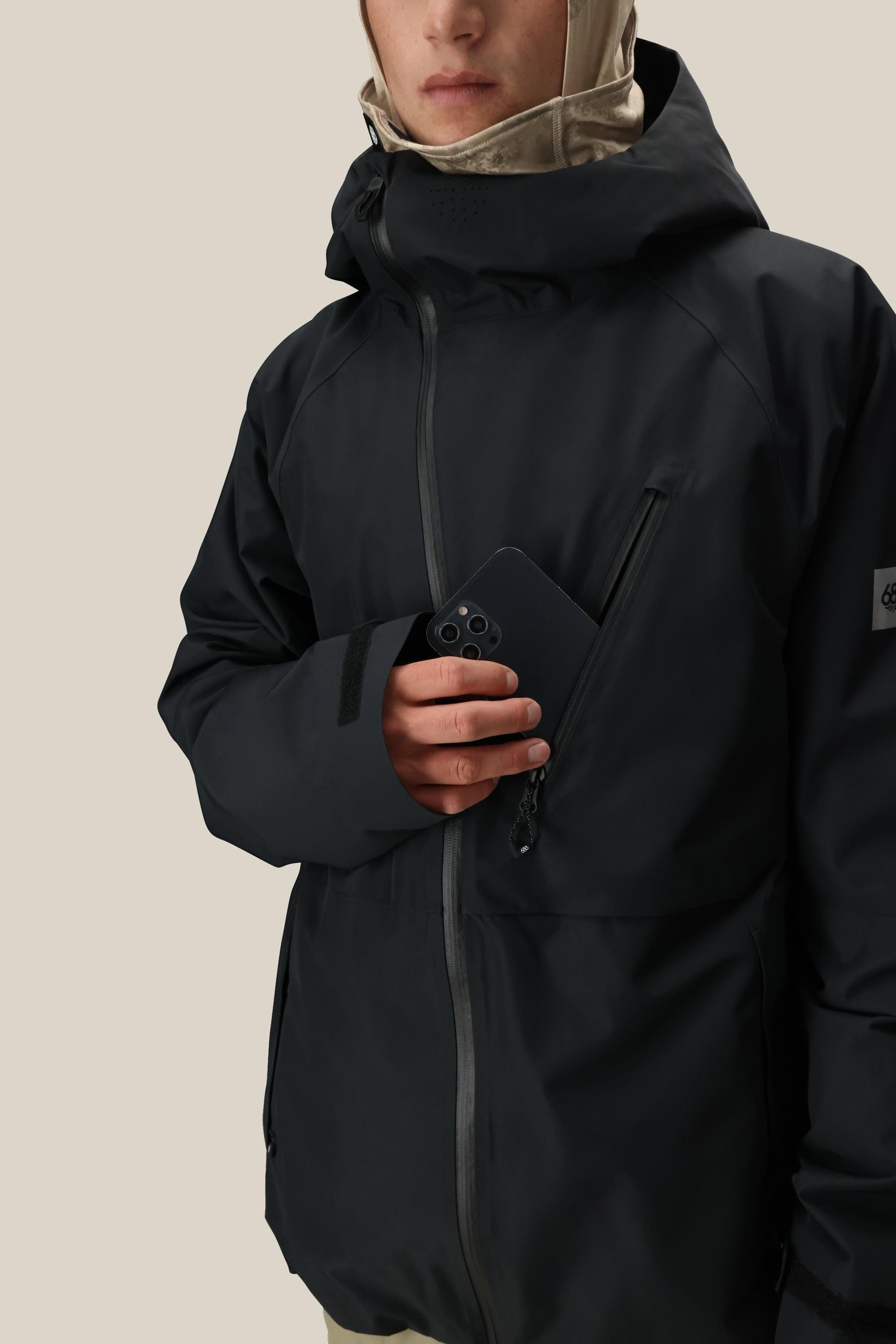 A person wearing the 686 Men’s Hydra™ Thermagraph® Jacket in black and a light-colored balaclava holds a black smartphone partially tucked into the jacket’s chest pocket, set against a neutral background.