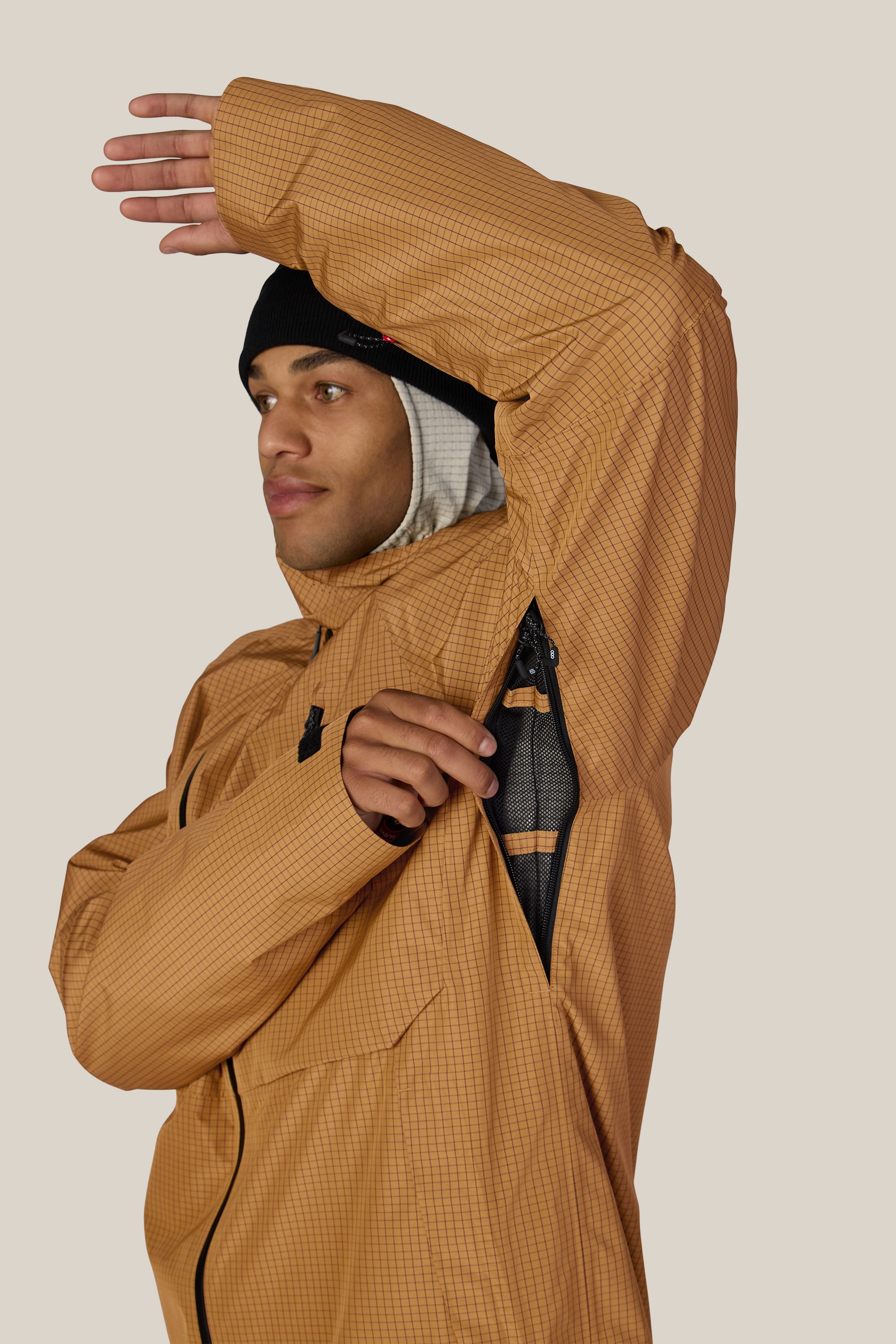 A person wearing the 686 Men’s Hydra™ Thermagraph® Jacket in a tan checkered pattern and a black beanie lifts their arm to unzip the underarm vent, revealing mesh fabric beneath. The background is plain and light-colored.